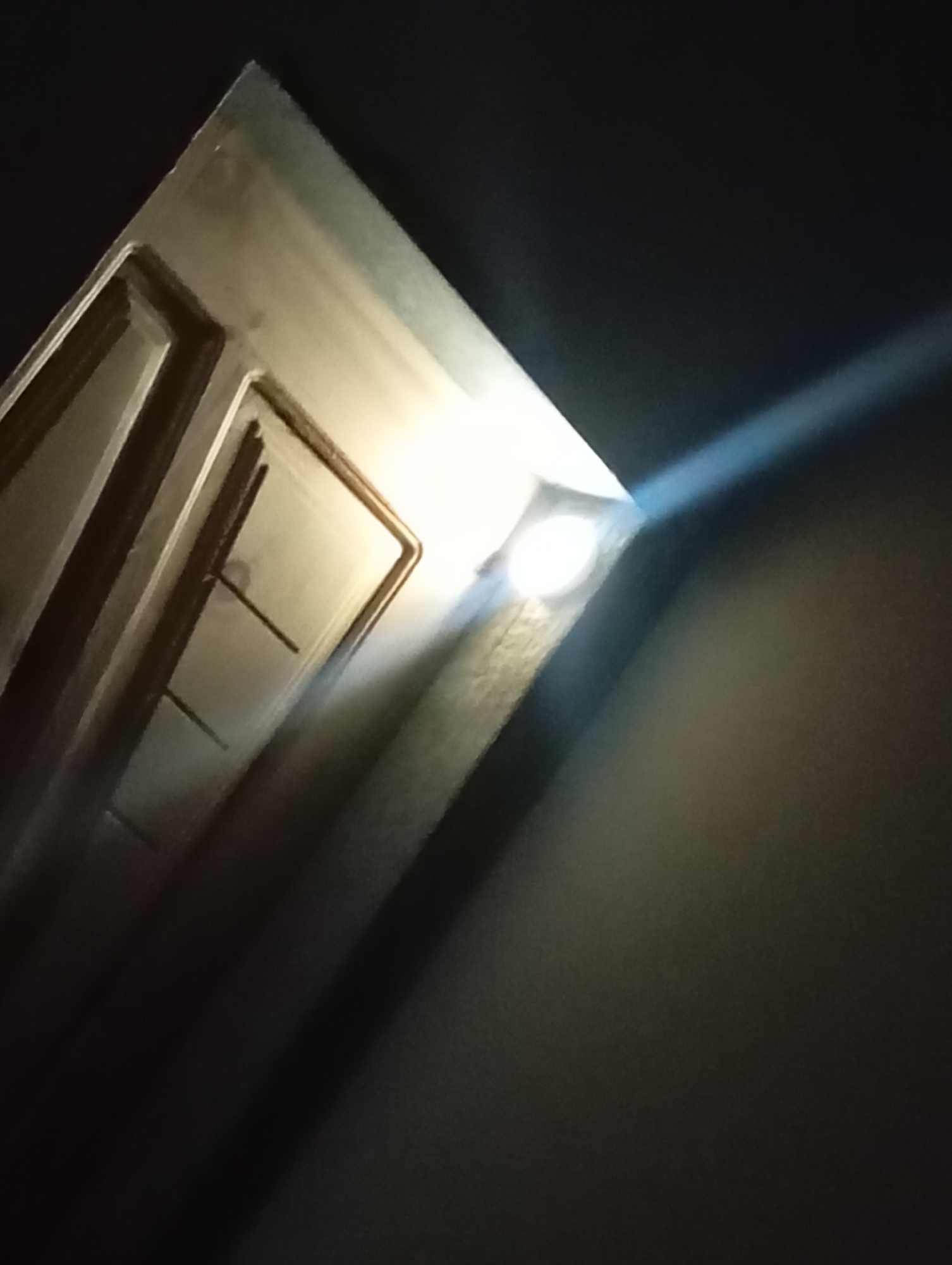 A low-angle, indoor shot captures the top corner of a cream-colored door against a dark background. A bright, white light emanates from a small, square fixture mounted in the corner where the door and wall meet, casting a distinct beam across the dark surface of the wall. The door appears to have a textured design with recessed panels.