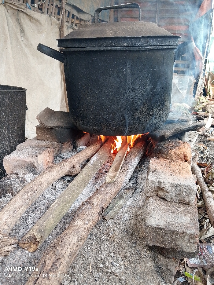 A large, black metal pot with a lid sits atop a small fire made of four wooden logs on a dirt and brick base. The pot has a handle on the left side. The fire is small and contained, with flames visible. The pot is sitting on four pieces of wood with a fire burning beneath it. The scene is taken outside, most likely in the backyard of a home. The scene is located at the coordinates -6.419, 107.978, which corresponds to a rural area in the city of Anjatan, Indonesia. There are no people visible in the image. The pot appears to be used for cooking. The image was taken on a phone, as evidenced by the watermark on the bottom right corner.