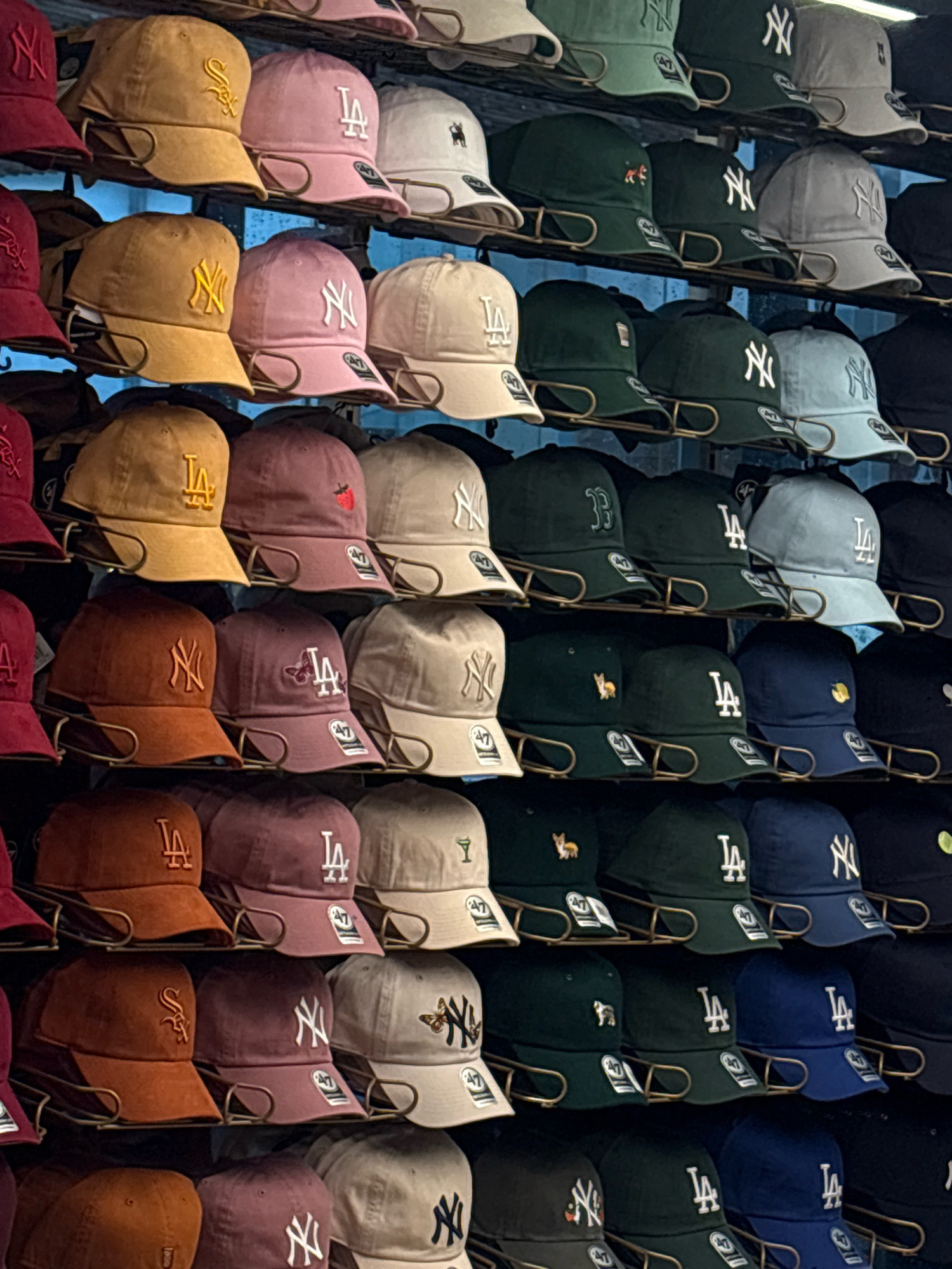 The image shows a wall of baseball caps displayed on shelves in a retail setting. The caps are arranged in rows and columns, with a variety of colors and logos. Some of the visible logos include "NY" (New York Yankees), "LA" (Los Angeles Dodgers), and what appears to be a strawberry. The caps are predominantly in shades of tan, pink, burgundy, green, and blue. The overall environment suggests a store specializing in sports merchandise. There are no people or discernible city landmarks visible, and therefore, no specific location within Mexico City can be identified from the image alone.