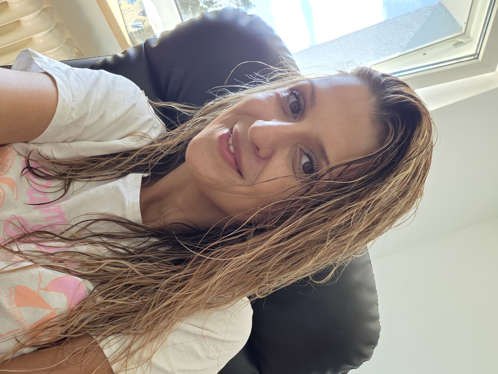 A woman with wet, blonde hair is shown in a selfie taken indoors. She is sitting in a black leather chair in front of a window. The woman is wearing a white t-shirt with a pink and orange graphic design. Her hair is wet and appears to be styled down, falling around her shoulders. She is looking at the camera and smiling. The background shows a window with natural light coming through, and a radiator is visible below the window. There is also a tissue box on a shelf next to the window. The overall setting appears to be a home or office environment. There is no indication that this image was taken in Munich, Germany.