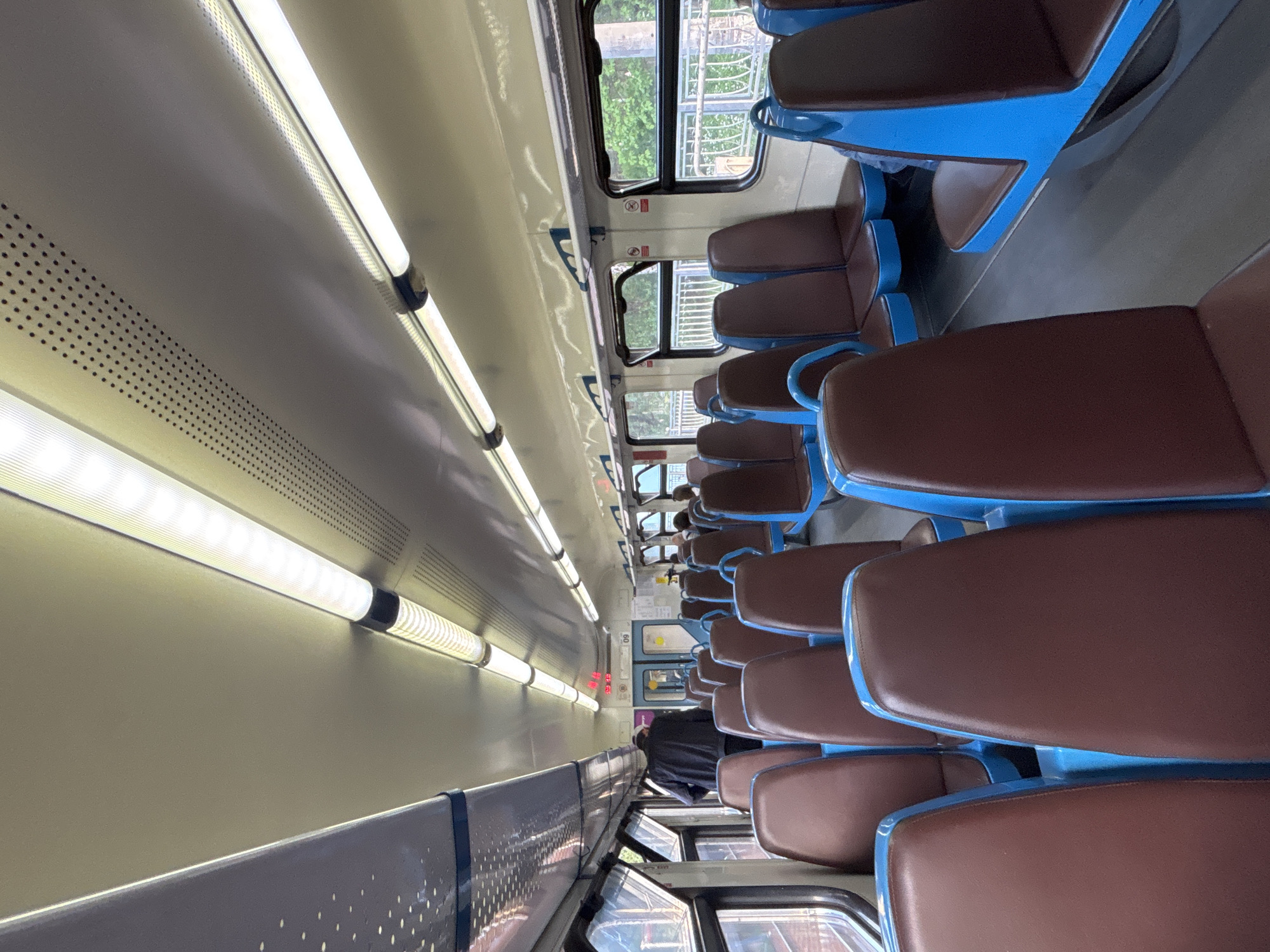 The image displays the interior of a commuter train car, likely in Komarovo, Russia, rotated 90 degrees clockwise. The scene captures a long aisle with rows of seats predominantly empty. The seats feature distinct brown upholstered cushions and bright blue metal frames or bases, arranged in pairs facing each other in some sections and forward-facing in others.
On the left side of the frame, serving as the "ceiling" in a normal orientation, are multiple long, bright fluorescent or LED tube lights that illuminate the car. These lights are mounted on a cream-colored panel system, which also features sections with small, uniform perforations, likely for ventilation or speakers.
The "floor" of the train car, seen on the right side of the image, has a light grey finish. Windows line the right side of the car, offering glimpses of green foliage and a metallic fence or railing outside, suggesting the train is either stopped at a station or moving through a natural landscape. Small red "No Smoking" symbols are visible on stickers near the windows.
Towards the far end of the car, a person dressed in dark clothing is partially visible, either sitting or standing. Above the far door, a digital display shows red numbers, possibly "23:13" and "09", which could indicate time or other train information. The overall impression is one of a clean, well-lit, and functional public transport space during what appears to be daytime, given the natural light from the windows.