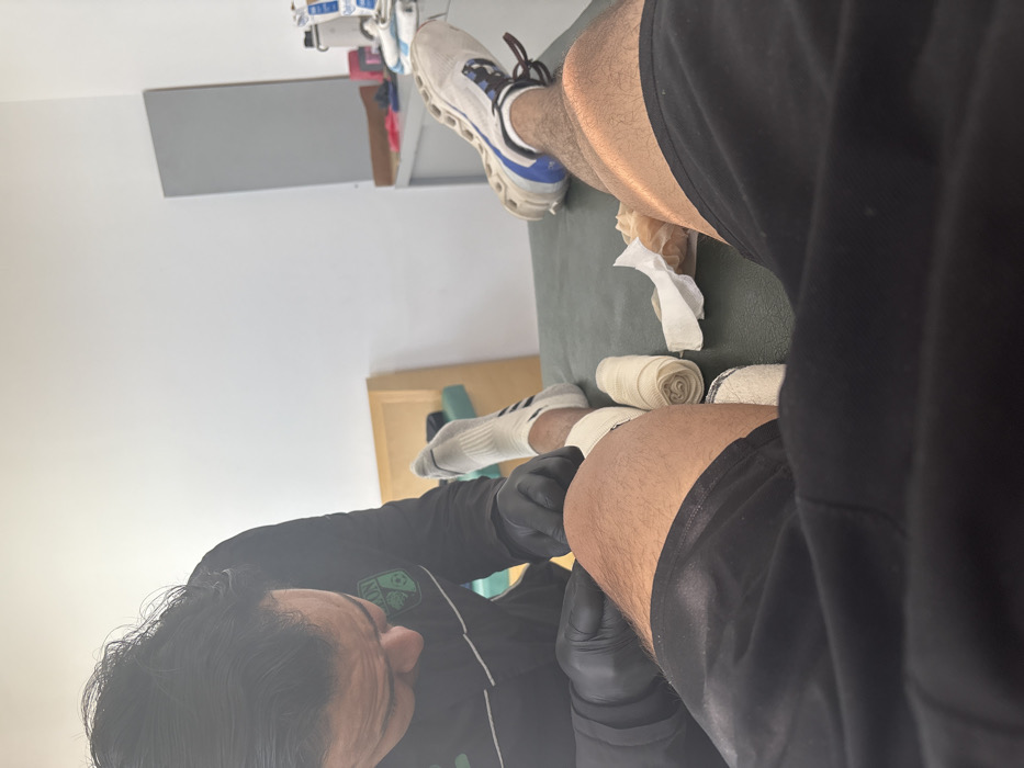 A person with black gloves is wrapping bandages on their leg, which is resting on a green surface. Beside the wrapped area are two rolls of bandages. Above, another leg wearing a sock and shoe sits on the same surface. The person wrapping the bandage is wearing a black and green athletic shirt. Tissue paper is scattered on the surface near the wrapped legs and bandages. A shelf with various items is visible in the background.