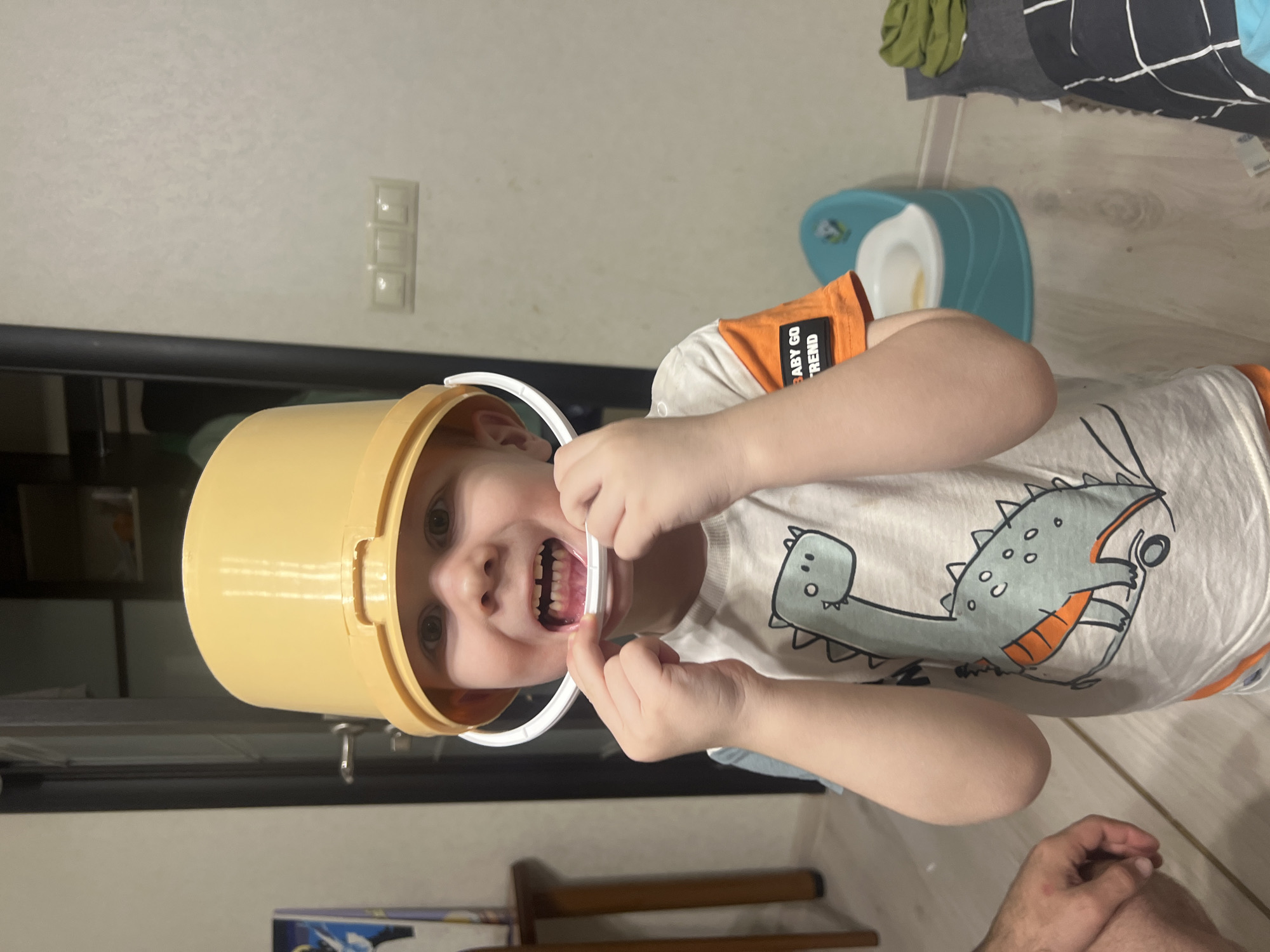 A boy is holding a white plastic handle in front of his open mouth, grinning, and wearing a yellow bucket on his head. He is wearing a white T-shirt with a gray dinosaur printed on it. There is a child's potty chair visible in the background. The location is identified as the city of Chaikovsky, Russia.