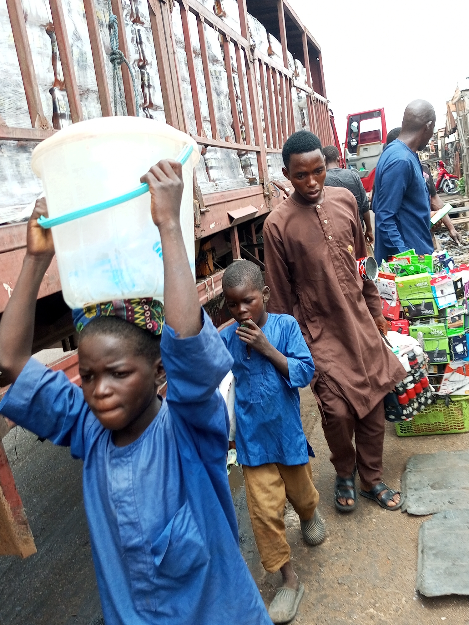 The image depicts a scene in the city of Gombe, Nigeria, likely a market or commercial area. In the foreground, a young boy wearing a blue robe and tan pants carries a large, clear plastic bucket filled with water on his head. He has a colorful cloth wrapped around his head beneath the bucket. To his right, another boy, also in a blue robe, is eating something. Behind them, a man in a brown, buttoned robe walks alongside a stall laden with various goods, including boxed items and smaller containers. Another man in a blue garment stands further back, looking towards the right. The background reveals a large, red truck with a metal cargo area, suggesting transportation and logistics are part of the environment. The ground appears to be dirt or a rough paved surface, with some debris and objects scattered around. The overall atmosphere seems bustling and busy, characteristic of a Nigerian market.