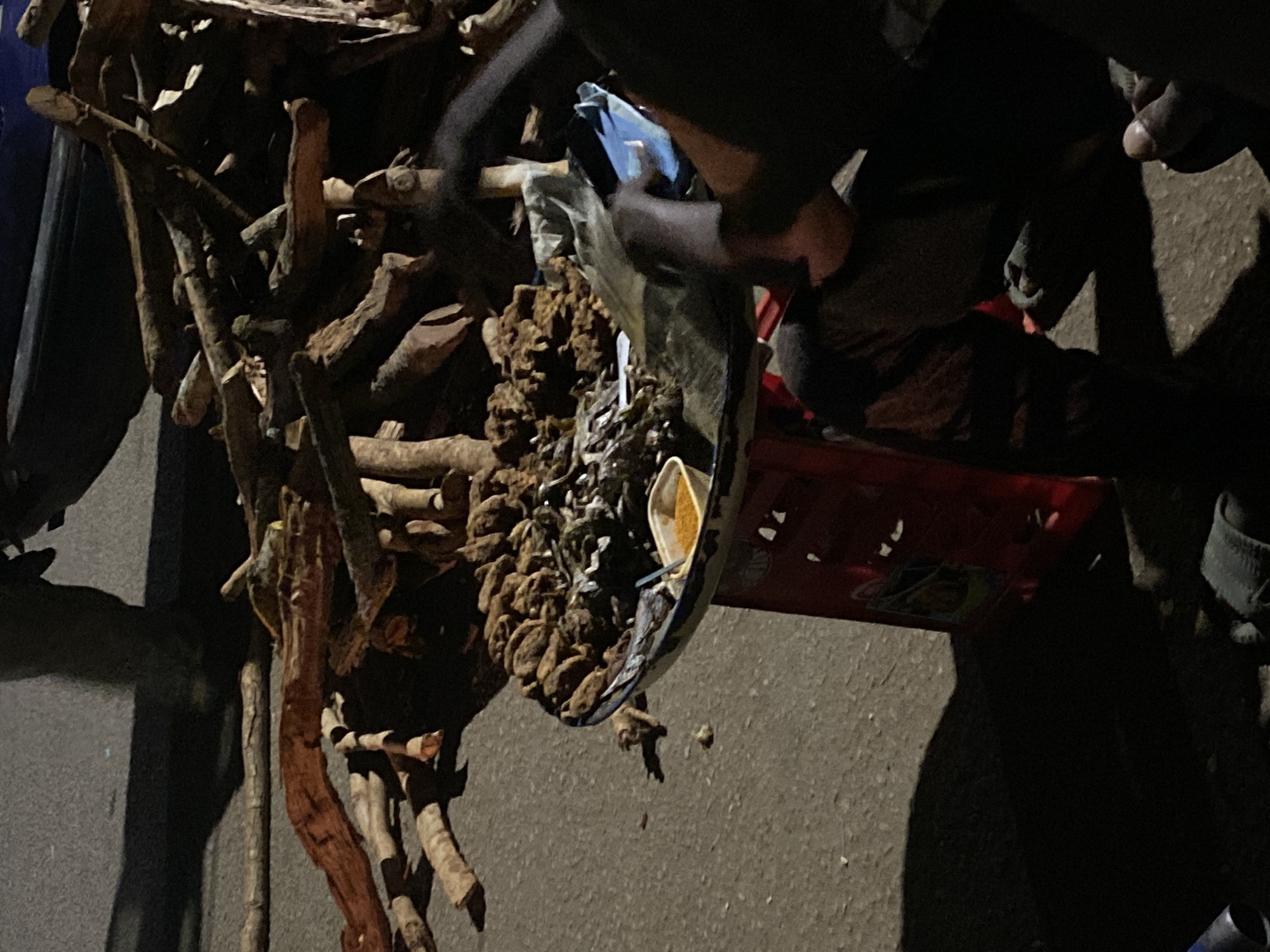 A low-angle shot depicts a street scene in Bukuru, Nigeria, where someone is seen holding a blue-rimmed, white and gold container filled with a mixture of brown, knotted items (possibly foodstuff), white bits and pieces, and yellow powder. The container is angled downward. To the left, a collection of branches and sticks is visible. The ground surface is asphalt. The lighting suggests the time of day is dusk or night. A red object is located on the right. A person can be seen in the background. The photograph is taken close to the subjects and objects in view.