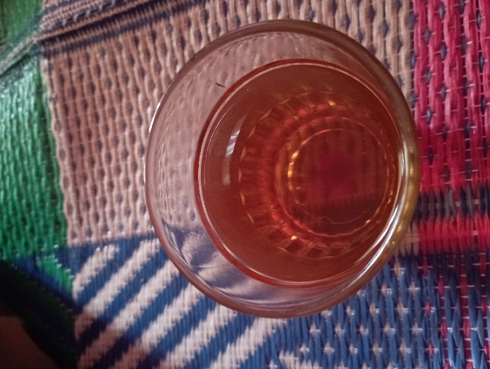 An overhead shot captures a clear glass containing an amber-colored liquid, resting on a multi-colored woven mat. The glass is positioned centrally, slightly towards the right, with light reflecting off the liquid's surface and the glass rim. Concentric circular patterns are visible at the bottom of the glass through the liquid.

The woven mat, likely made from synthetic material or straw, features distinct sections of color and pattern. On the far left, a green section is partially visible. Adjacent to it is a wider section characterized by light beige/off-white stripes interwoven with blue, creating a textured pattern. To the right, beneath and beside the glass, a prominent section displays red and dark blue/black threads woven into a crosshatch or checkerboard pattern. The intricate weaving of the mat is a notable detail, providing a textured background for the glass.

The scene is static, depicting an object in its setting rather than an activity. The lighting suggests an indoor or shaded outdoor environment. The type of woven mat is common in Southeast Asian households, aligning with the Pubian, Indonesia context. No people are visible, and no text is present in the image. The overall impression is a simple, still-life composition.
