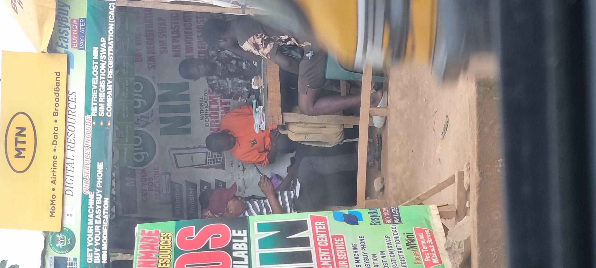 A street-level medium shot shows a small outdoor stall in Otta, Nigeria, likely serving as a mobile money and business services hub. The stall is adorned with colorful banners from companies like MTN, Glo, and EasyBuy. In the foreground, a blurred yellow object, possibly part of a vehicle, partially obscures the view.
Behind a wooden table, three individuals are engaged in activities related to the stall's services. A man in a striped shirt and a maroon cap is seated, looking down at his phone. To his right, a man in an orange t-shirt is also focused on his phone, with a stack of papers on the table in front of him. Further to the right, another man, wearing a camouflage-patterned shirt, appears to be attending to business, with cash and documents on the table. The presence of a light-colored backpack suggests mobility or the personal belongings of one of the individuals.
The background of the stall features various signs and graphics related to mobile network services, including "DIGITAL RESOURCES," "GET YOUR MACHINE," "BUY YOUR EASYBUY PHONE," "SIM REGISTRATION/SWAP," and "COMPANY REGISTRATION (CAC)." The ground surface is dusty, consistent with an outdoor environment in Nigeria. The overall scene depicts a typical small business operating on a roadside in a Nigerian city.