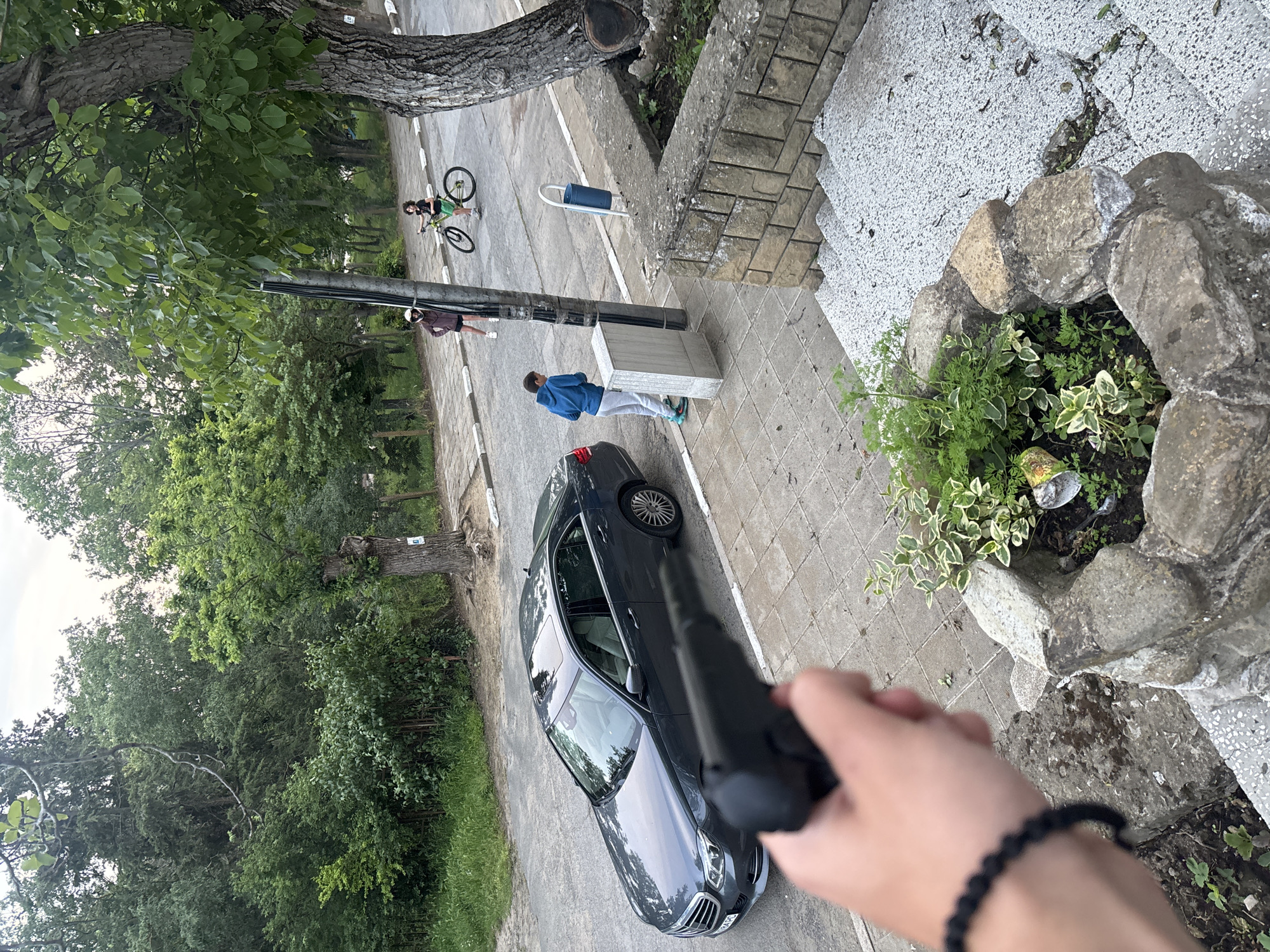 The image, taken from an elevated position, shows a street scene with a parked dark-colored car. A person in a blue jacket and grey pants stands next to a light-colored structure on the sidewalk. Further down the street, another individual is walking while a third person is riding a bicycle. A hand holding a dark object, possibly a gun, is visible in the foreground, obscuring part of the car. The setting appears to be in a residential area with trees and greenery visible. Based on the prompt, the location is in Goritsa, Bulgaria.