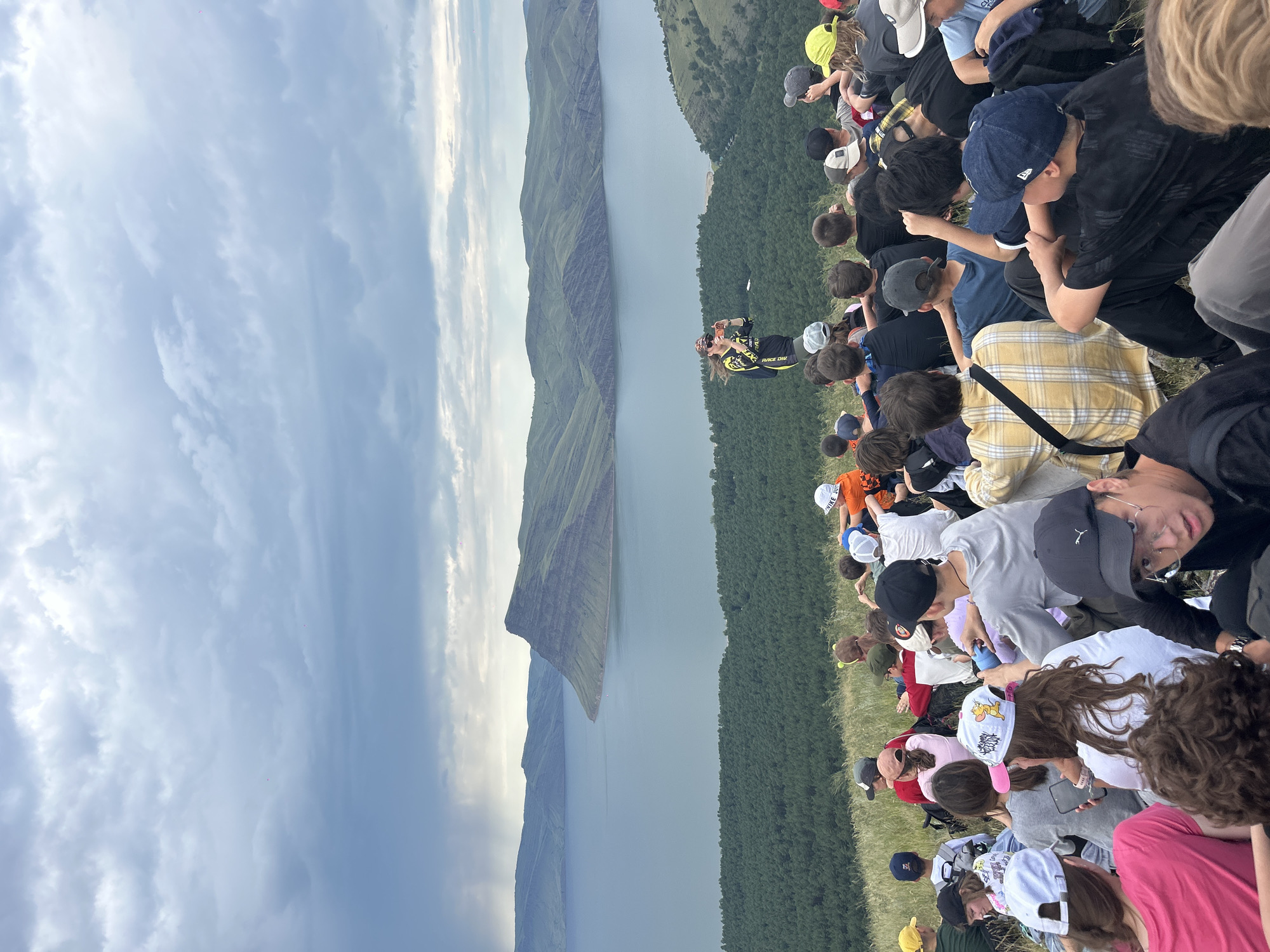 In a landscape that appears to be the vicinity of Nikolo-Petrovka, Russia, a large group of young people are gathered on a grassy hillside, facing a scenic view of a body of water and distant, verdant mountains. The sky above is overcast with dramatic, grey clouds. In the mid-ground, a lone individual, possibly an instructor or guide, stands facing the group, holding what appears to be a phone, possibly taking a picture or recording. The group is seated on the slope, some individuals are looking towards the instructor, while others are looking out at the view. Their attire is casual and varied, suggesting an outdoor excursion or educational activity. The overall impression is one of a shared experience in a natural setting.