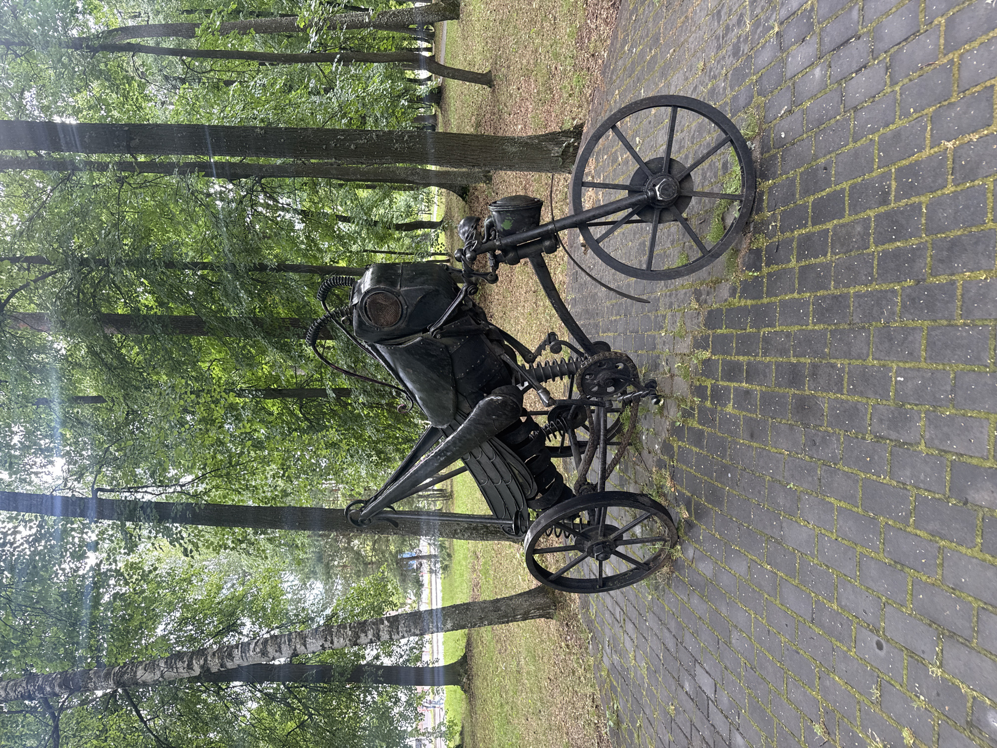 This media file shows a sculpture of a diver's helmet and suit mounted on a three-wheeled vehicle, possibly a custom-made bicycle or a small, wheeled platform. The sculpture is made of dark metal and features intricate mechanical details, including springs and articulated parts. The diver's helmet has a distinct, round faceplate with mesh detailing. The vehicle has two large spoked wheels at the rear and a smaller wheel at the front.
The sculpture is situated outdoors on a paved walkway made of dark rectangular bricks with moss growing in the gaps between them. The setting is a park or wooded area, with numerous trees visible in the background, their leaves creating a lush green canopy. Sunlight filters through the leaves, casting dappled light. The grass in the background is green and appears well-maintained. The overall scene suggests a public space, possibly an art installation in a park.
There is no visible text in the image, and the time of day appears to be daytime, likely morning or afternoon, given the lighting conditions. The weather seems pleasant, with clear skies and sunlight. The image evokes a sense of artistry and perhaps a touch of whimsy, given the unusual combination of a diver's suit and a wheeled vehicle. The location context provided is Sarov, Russia, suggesting this is an outdoor exhibit in that city.