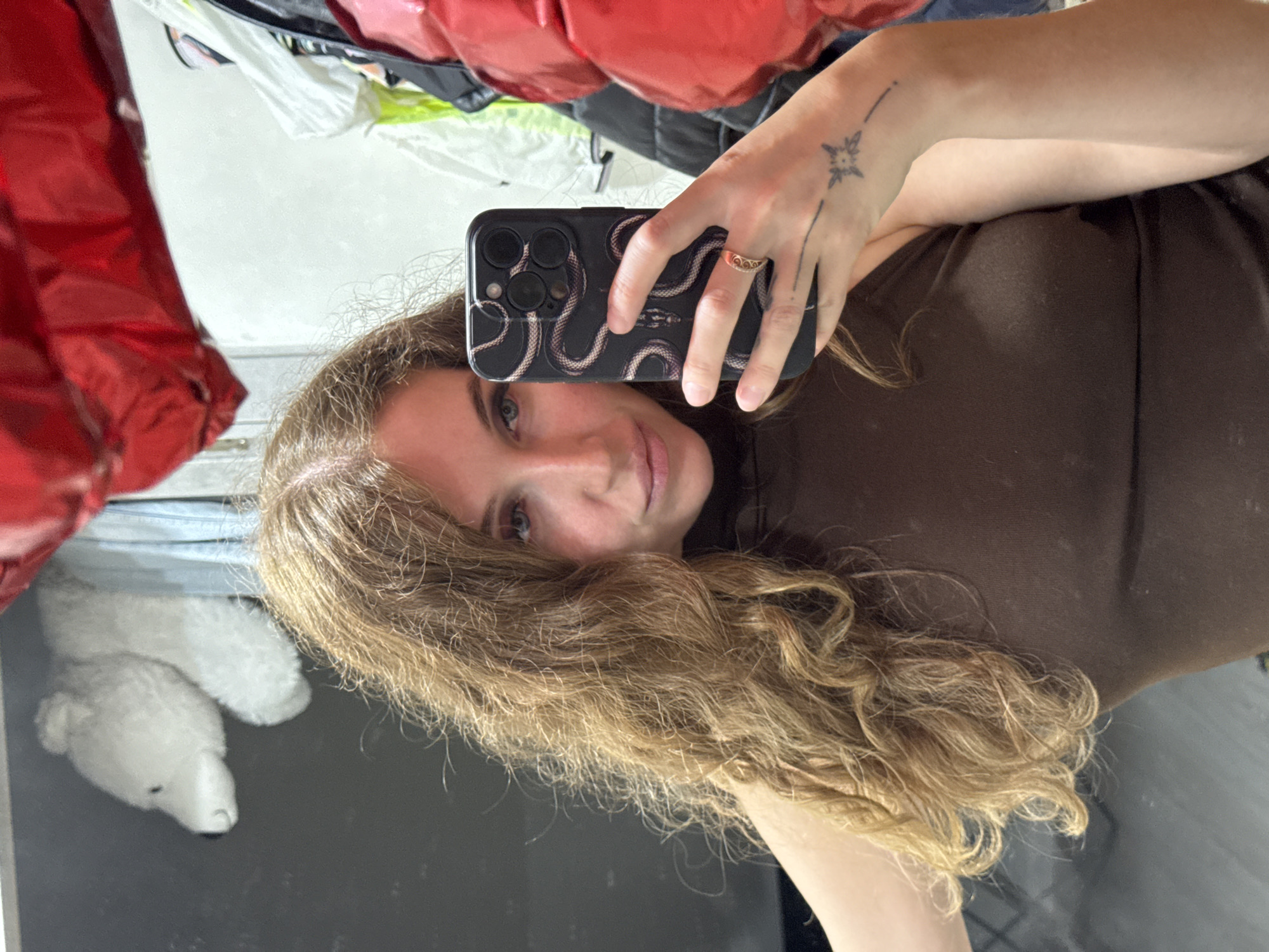 A woman with long, wavy blonde hair is taking a mirror selfie. She is wearing a brown sleeveless top. She is holding a black smartphone with a snake design on its case. On her right hand, she is wearing a ring and has a tattoo on her wrist. In the background, reflected in the mirror, are clothing items, including a red puffer jacket and a white stuffed bear. The setting appears to be indoors, possibly a closet or dressing area. There is no identifiable information that places this image in Chaikovsky, Russia.