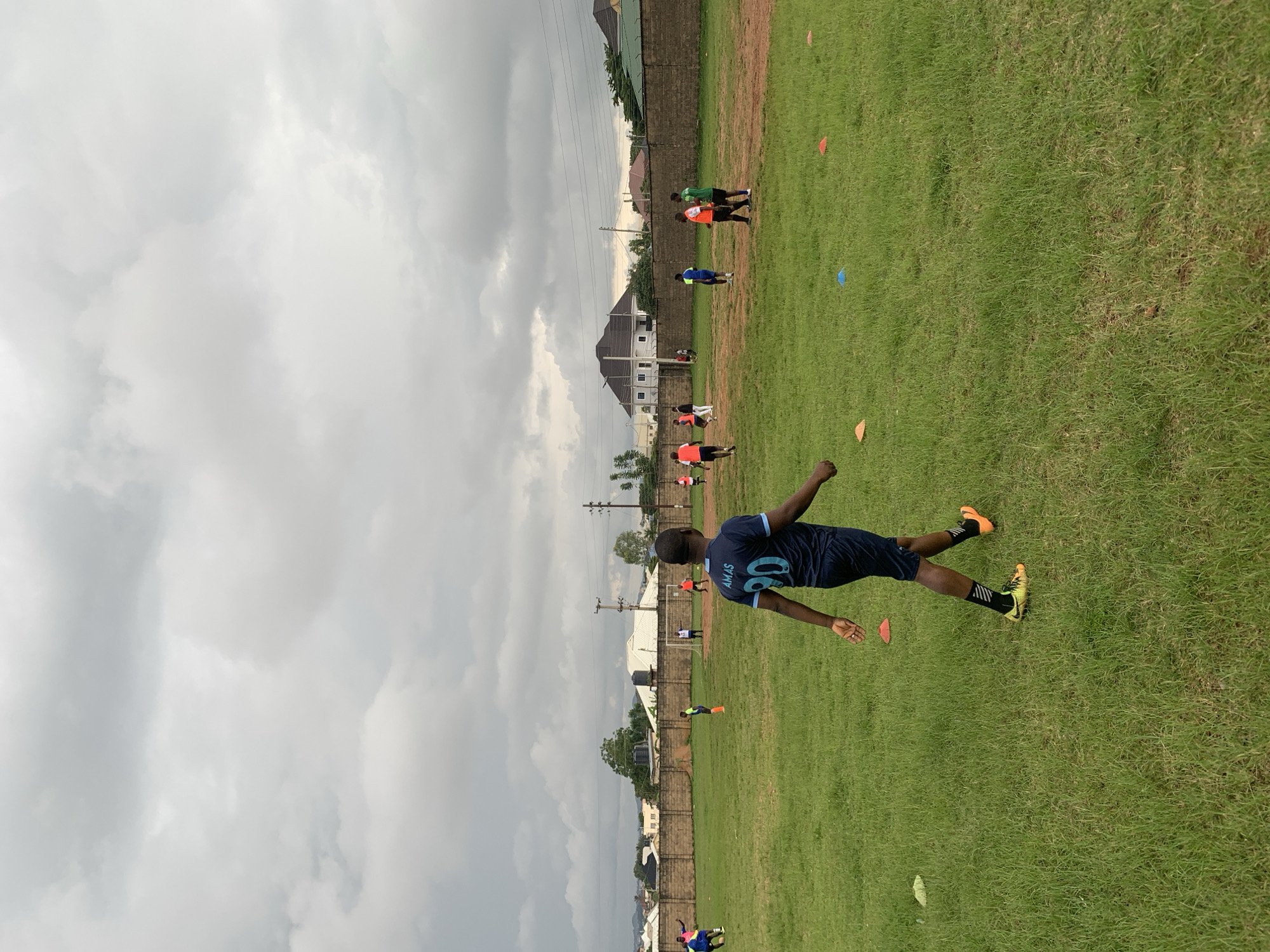 A group of young men in athletic wear are engaged in a soccer training session on a grassy field. The sky is overcast with heavy clouds, suggesting either early morning or an impending storm. In the foreground, a young man wearing a dark blue jersey with the inscription "AMAS 90" and dark shorts is walking away from the camera. He has bright yellow soccer cleats with orange accents and black socks. Several colorful cones, likely used for drills, are scattered across the field. In the background, other individuals are dispersed, some appearing to be practicing or observing. Beyond the field, a long, weathered brick wall encloses the area, with buildings and vegetation visible on the other side. The overall impression is of a casual, community-level sporting activity taking place in an urban or semi-urban environment. The location is identified as Jos, Nigeria.