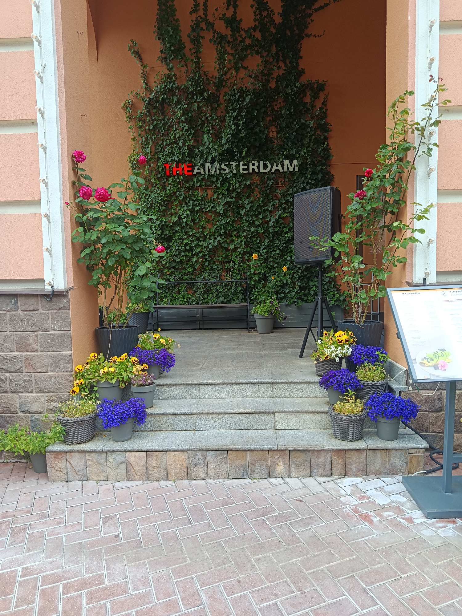 The image shows the entrance to a restaurant named "THE AMSTERDAM" in Estosadok, Russia. The entrance is decorated with lush green ivy covering the wall and a large rose bush with red roses in front of it. Several potted plants with colorful flowers, including blue and yellow pansies, are arranged on the steps leading up to the entrance. A speaker on a stand is positioned to the left of the entrance, and a menu stand is to the right. The building's exterior features a textured orange wall and stone accents. The foreground shows a paved walkway with a herringbone brick pattern.