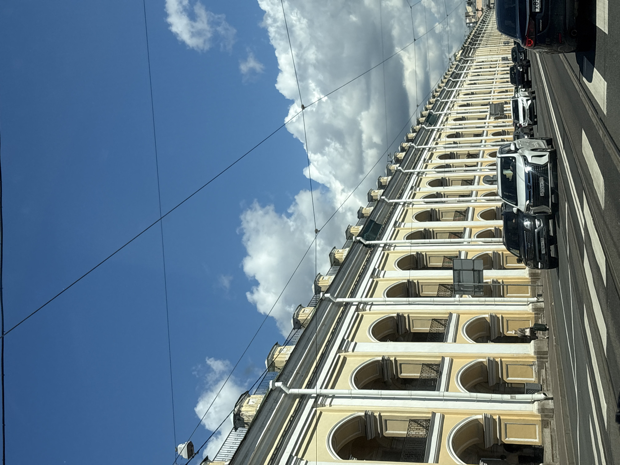 This is a street-level, eye-level, medium shot of a long, yellow building with an arched colonnade, likely in Saint Petersburg, Russia, given the architectural style. The building features multiple stories with repeating arched openings, many of which have balconies. White decorative elements and downspouts accent the yellow facade. Overhead, electrical wires crisscross the bright blue sky, which is dotted with fluffy white clouds.
The street in front of the building is a multi-lane road with white crosswalks. Several cars are driving on the road, including a dark-colored SUV on the right, a white minivan in the center, and other vehicles in the distance. One person, a woman carrying luggage, is walking on the sidewalk in front of the building. The overall scene depicts a sunny day in a historic urban environment.