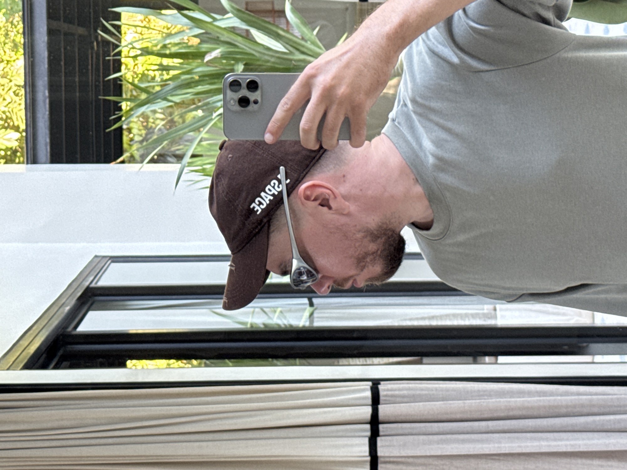 This image captures a person from the chest up, rotated 90 degrees clockwise, giving the appearance of someone looking downwards to their left. The subject is likely taking a selfie or a photo of their reflection, holding a modern smartphone (identifiable by its triple camera module) with their right hand positioned above their head.
The person wears a light grey short-sleeved t-shirt and a brown baseball cap. On the front of the cap, mirrored white text is visible, which reads "SPACE" when un-mirrored. They also have stubble or a short beard and are wearing silver-framed glasses or sunglasses that are pushed up onto the bridge of their nose.
The background, visible around the person, features bright green, dense foliage, suggesting an outdoor or semi-outdoor tropical setting. There is a white wall or structure behind them, and part of a dark-framed window or door frame is visible. Below this, light-colored horizontal blinds or louvers can be seen. The bright lighting indicates it is daytime. The scene evokes a warm climate, consistent with the provided location context of Kuta Selatan, Indonesia.