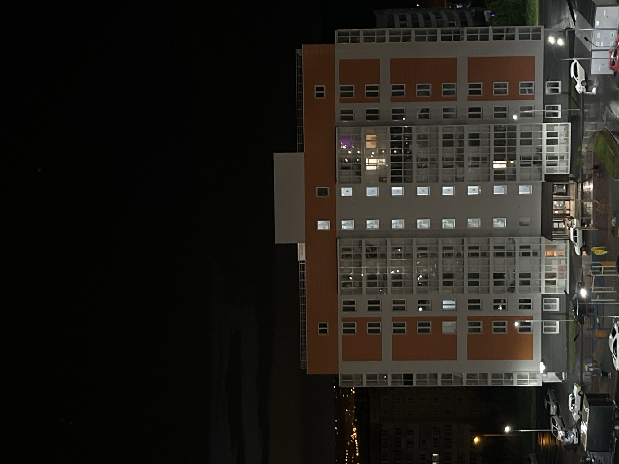 The media file captures a nighttime view of a modern, multi-story building, likely an apartment complex or dormitory, situated in Krasnoyarsk, Russia. The building's facade features a striking design with orange and white sections, interspersed with numerous windows that are lit from within, suggesting occupancy. A prominent section of the building is dominated by large, grid-like windows, revealing interior spaces and reflections. Adjacent to the main structure, a smaller white building section is visible. The scene is set against a dark, almost black sky, indicating it is nighttime. The ground level shows a paved area with parked cars and streetlights casting illumination. One car appears to be white, and another is red, while several other vehicles are visible but less distinct. The wet pavement suggests recent rain, contributing to the reflections of lights on the ground. The overall mood is quiet and still, typical of a residential area at night. No people are clearly discernible, but the lit windows imply their presence within. There is no visible text in the image.