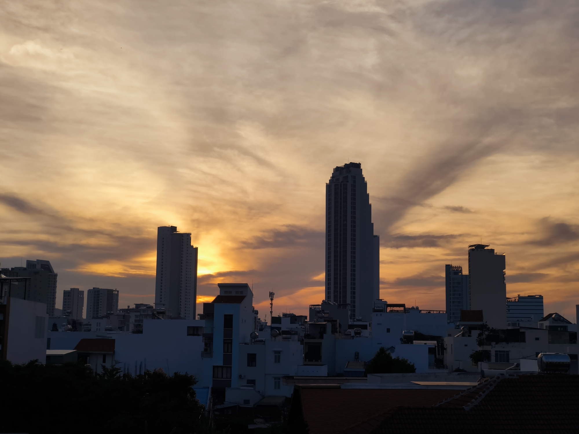 The image depicts a cityscape at sunset, likely in Nha Trang, Vietnam, characterized by a dramatic sky filled with orange and yellow clouds. Silhouettes of various buildings, including several tall high-rise structures and smaller residential or commercial buildings, dominate the midground and foreground. The sky exhibits a sunburst effect with rays extending outwards from behind the clouds, creating a vibrant and atmospheric scene. The lower portion of the image is darker, showing the rooftops and structures of the city in shadow. There are no visible people or active events, presenting a still, architectural panorama.