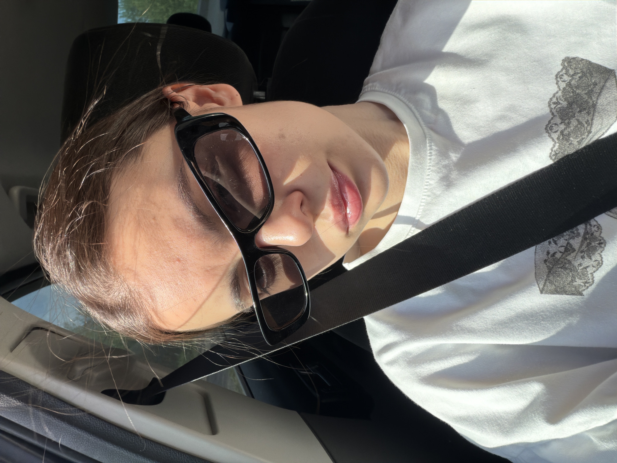 A person wearing sunglasses and a seatbelt is sitting in a car. The person is wearing a white t-shirt. The location is unknown. The image provides no information about the city of Chaikovsky, Russia.