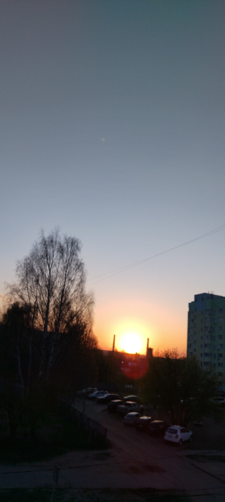 The image depicts a sunset in Chaikovsky, Russia, likely taken from an elevated position. The sky transitions from a pale blue at the top to a vibrant orange near the horizon where the sun is setting. Silhouetted trees stand on the left side of the frame. To the right, a multi-story building is visible. Below the horizon line, a parking area contains several parked cars. Utility lines are strung across the sky.