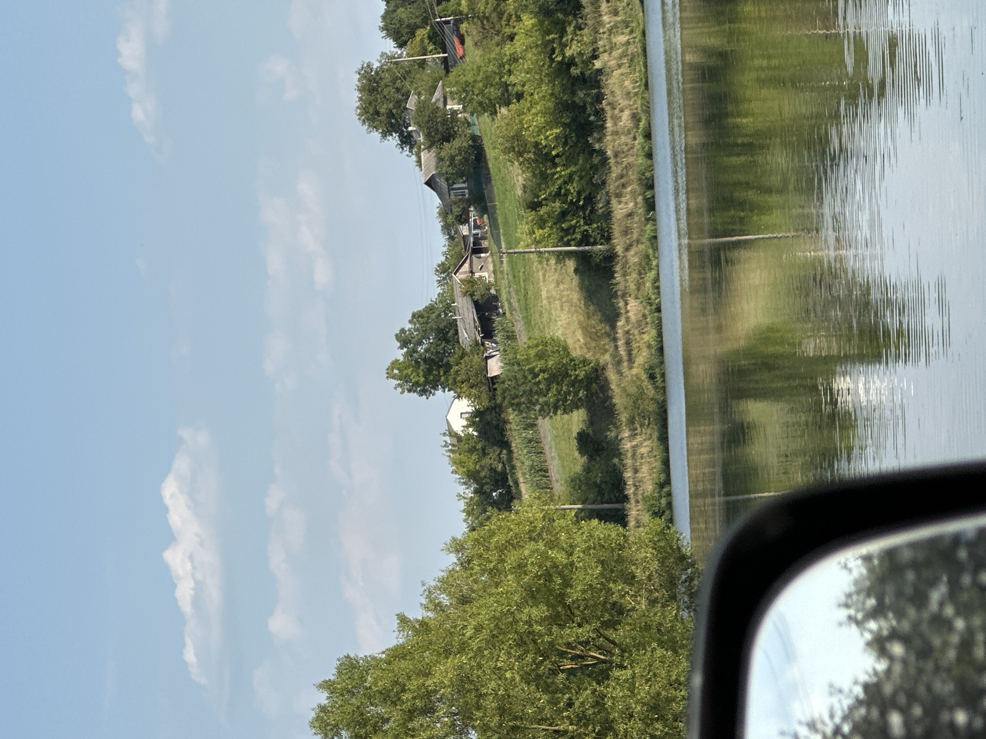 The image depicts a rural landscape, likely in Makiv, Ukraine. In the foreground, the reflection of trees and the sky is visible on the surface of a body of water, possibly a river or lake. A side-view mirror of a vehicle is partially visible in the lower left corner, reflecting more trees and the sky. The middle ground features a grassy embankment rising to a gentle hill. A few houses with sloped roofs are situated on the hillside, surrounded by trees and some cultivated fields, possibly corn. The background shows a clear blue sky with scattered white clouds. The overall scene conveys a peaceful, sunlit rural environment.