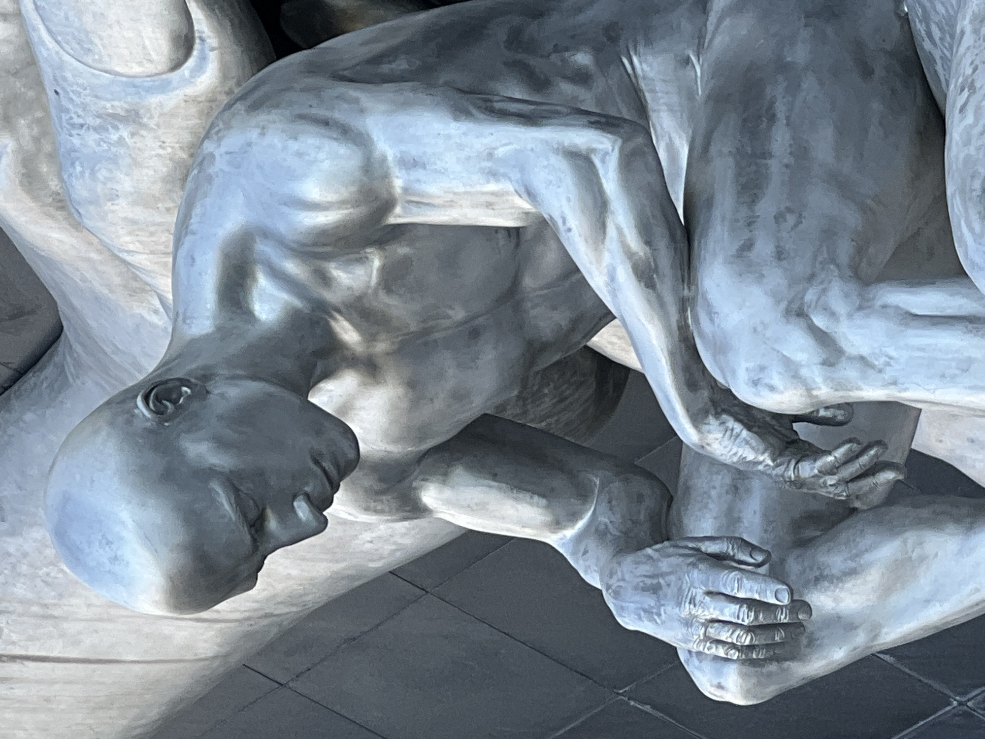 This is a close-up, eye-level, medium shot of a metallic sculpture depicting a bald, muscular male figure in a pensive pose. The figure is seated, with its head bowed and eyes looking downward. Its arms are resting on its knees, with hands delicately placed. The sculpture has a silver-toned, polished surface that reflects ambient light, creating highlights and shadows that emphasize the musculature and form of the body. The background is out of focus, with a textured, possibly tiled, dark surface on the left and smoother, lighter surfaces of what appear to be other parts of the same or a related sculpture. The overall impression is one of contemplation and introspection. The image does not contain information about the city of Krasnodar, Russia.