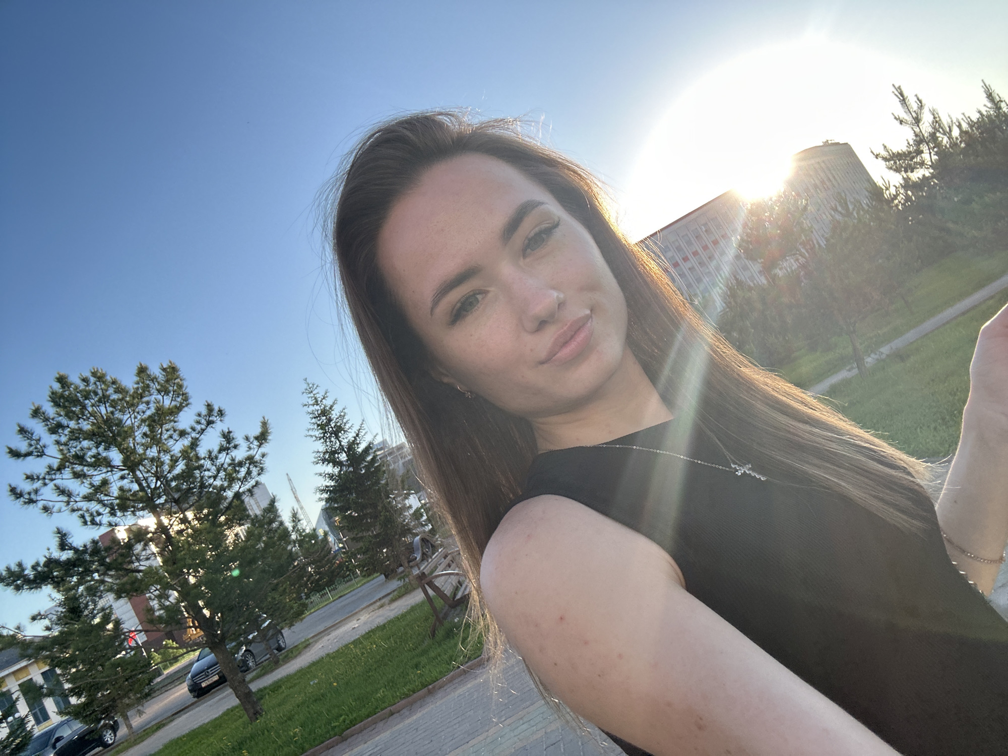 A young woman is featured in a close-up selfie taken outdoors during the late afternoon or early evening. She is positioned centrally, looking directly at the camera with a slight, calm smile or smirk. She has long, dark brown hair that catches the sunlight, green or hazel eyes, and fair skin. She wears a dark, sleeveless top and a delicate silver necklace with a small pendant.
The background is bathed in the warm glow of a low-setting sun, which creates strong backlighting and prominent lens flare, particularly behind the woman's head and to her right. The sky is a clear, bright blue.
The environment is an urban or suburban landscape. To the woman's left, several evergreen trees are visible against the blue sky, with a paved area and glimpses of vehicles further back. To her right and behind her, the sun shines brightly behind a large, distinct building characterized by a tall, cylindrical upper section and a lower structure with red and white horizontal stripes. More evergreen trees are present in this part of the background, along with green grass, paved sidewalks, and a road. Two dark-colored cars are parked or driving; one is a Mercedes-Benz sedan, whose front grille and a partial license plate (including "RUS" and some Cyrillic characters) are visible. A metal bench can also be seen. The scene conveys a bright, sunny day.