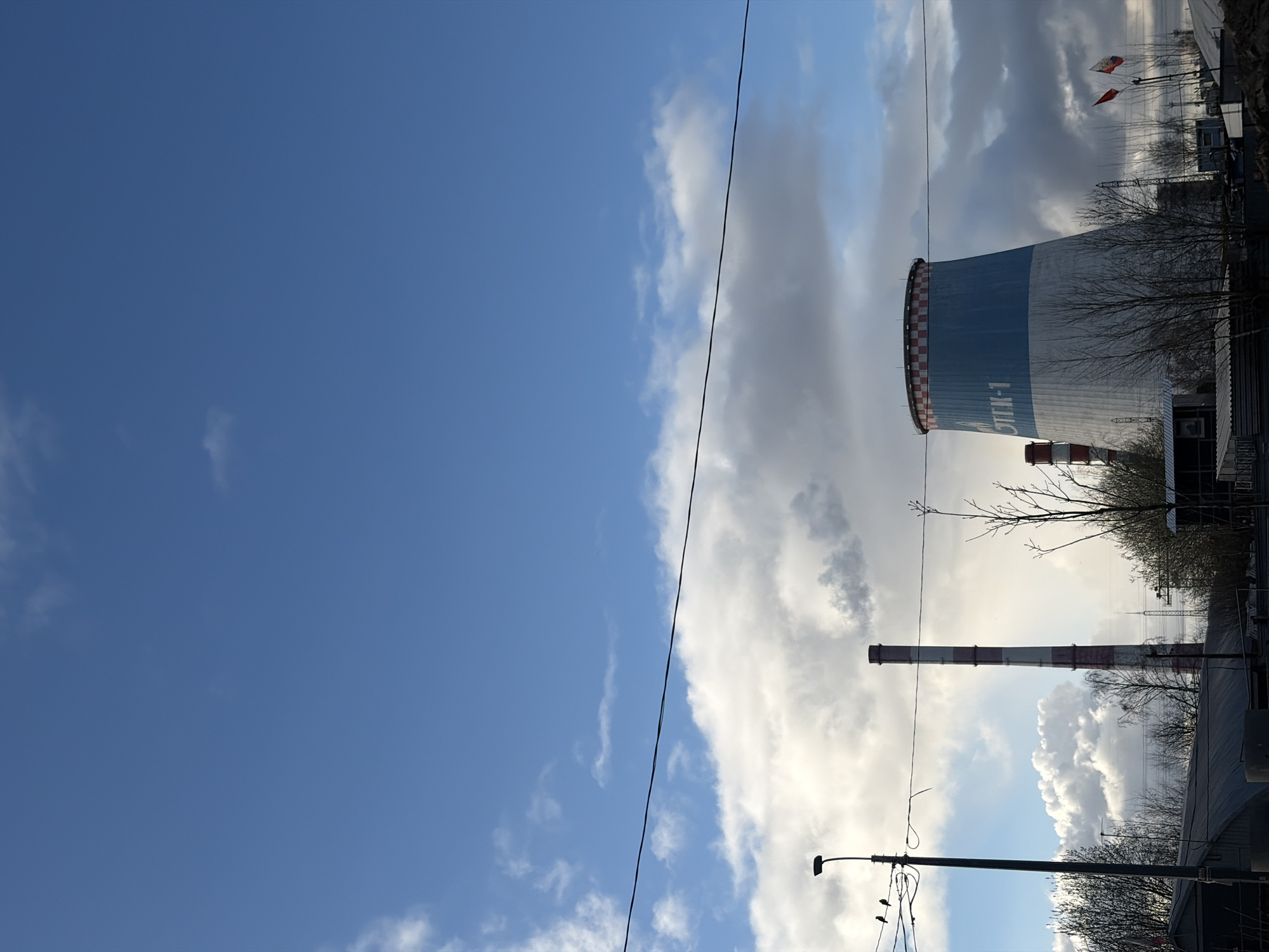 The image shows a view of the cloud-filled sky, next to a power plant in Saint Petersburg, Russia. Dominating the scene is a large cooling tower with the Russian text "CTTK-1" on its side. Several chimneys with red and white stripes stand nearby. Other structures, including what appears to be a power distribution area, can be seen in the background along with bare trees. A power line cuts across the sky, and a lamppost with some birds perched on it is visible in the lower portion of the picture.