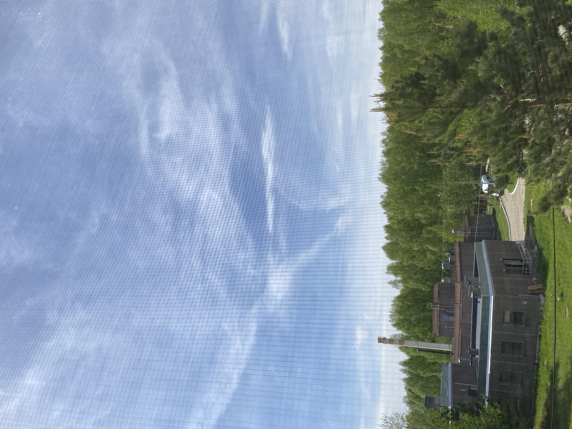 The image depicts a building complex with a tall chimney in the area of Марковский, Russia. The sky is visible with wispy clouds. The buildings appear to be made of a grey material and are surrounded by trees. A road and parked car are also visible in the background. The image seems to be taken through a mesh screen.