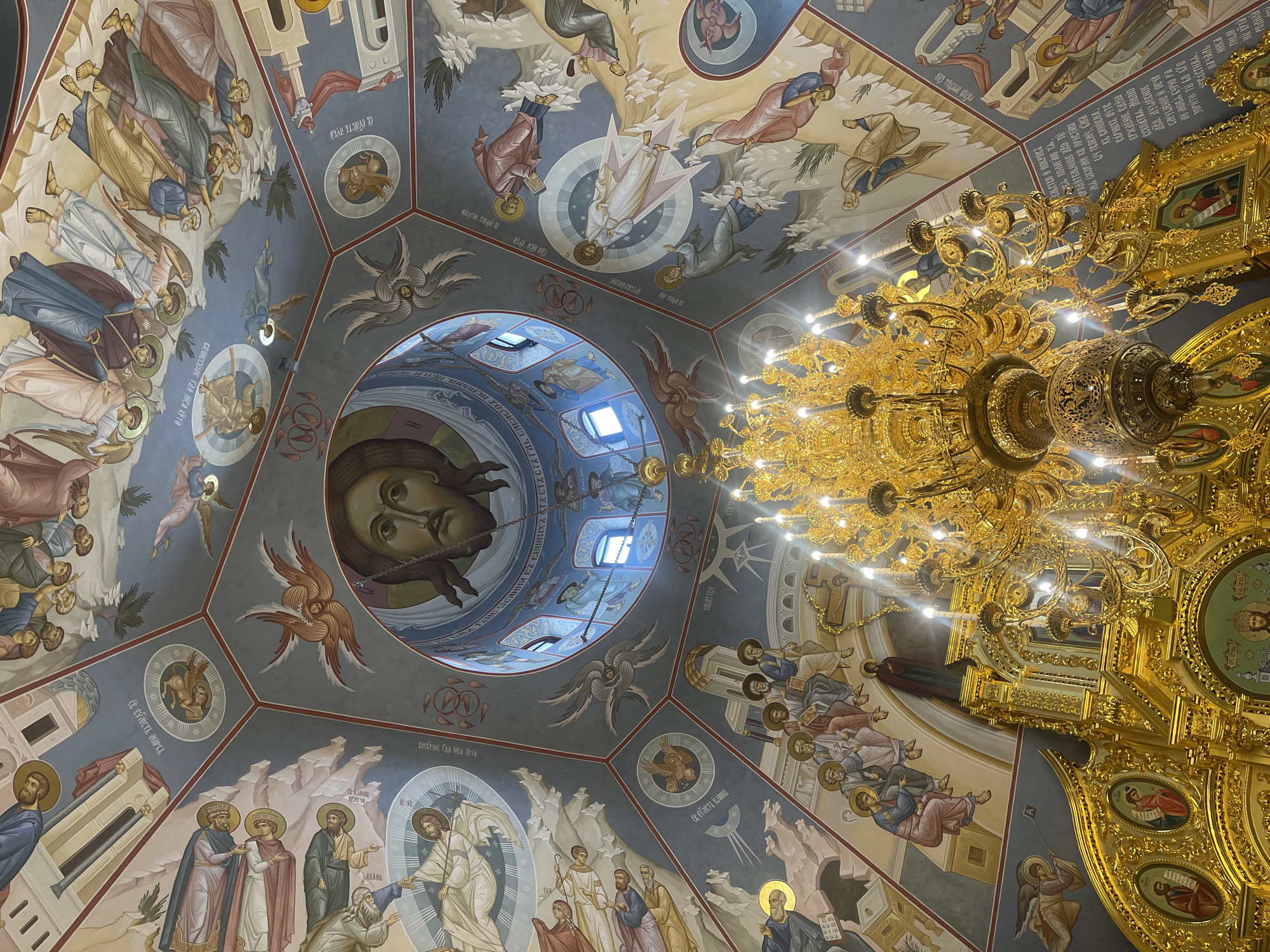 This is an interior view looking up at a church ceiling. The ceiling is adorned with religious frescoes, including portraits of figures and scenes featuring people in robes. The central dome features a close-up portrait of Jesus Christ. A large, ornate gold chandelier hangs from the ceiling, partially obscuring the artwork. The location of the image is in the city of Khimki, Russia.