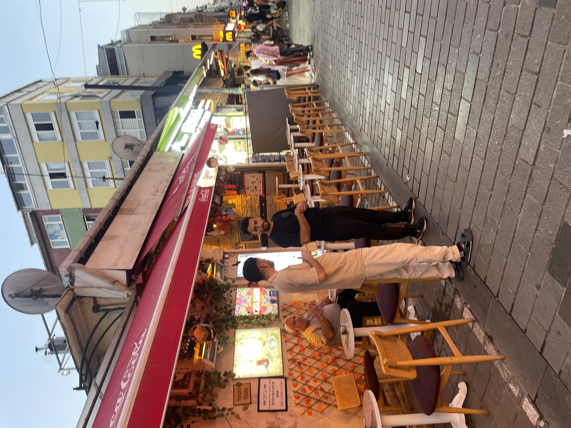 The image depicts a street scene in Kadıköy, Turkey, likely during the evening. A cafe with a red awning and Coca-Cola branding is prominent on the left, with outdoor seating arranged on a cobblestone sidewalk. Several individuals are visible, some seated at tables, while others are standing. Two men in the foreground appear to be interacting, one in a light-colored outfit and the other in black. In the background, a McDonald's sign is visible, suggesting the presence of commercial establishments. The architecture includes multi-story buildings with balconies and windows. The lighting suggests it is dusk or evening, with some artificial lights illuminating the cafe and street.