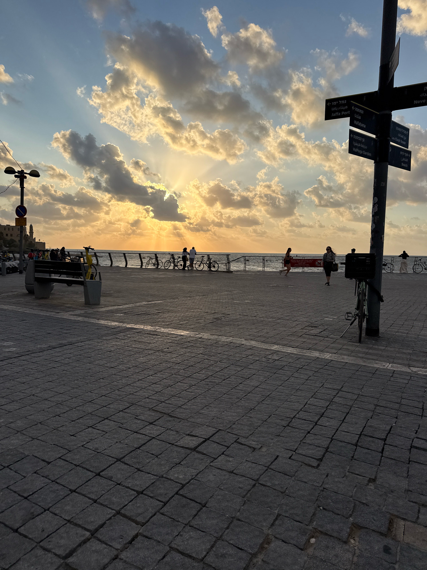 A wide-angle, eye-level shot captures a paved promenade overlooking the sea at sunset. The sky is filled with dramatic, sun-drenched clouds, with rays of sunlight breaking through. In the foreground, the ground is paved with dark, interlocking cobblestones, some of which appear worn or uneven.
To the left, a dark metal lamppost with a circular red and white sign attached to its base stands prominently. Further down the promenade, a concrete bench with two trash receptacles beside it is occupied by several people, silhouetted against the bright sky. A few yellow and black rental bicycles are parked near the bench.
Along the railing separating the promenade from the sea, various individuals are walking or standing. Several bicycles are also parked or leaning against the railing. The sea is calm, with a gentle wave pattern visible. In the distance, on the left side of the frame, a cluster of buildings, some with architectural features suggesting historical significance, are visible along the coastline, indicating the location is likely near Old Jaffa, Tel Aviv.
A tall black signpost with multiple directional signs is positioned on the right side of the promenade, further enhancing the urban, coastal atmosphere. A bicycle is leaning against this signpost. The overall scene conveys a sense of an active, public space enjoying the end of a day. The lighting suggests it is late afternoon or early evening.