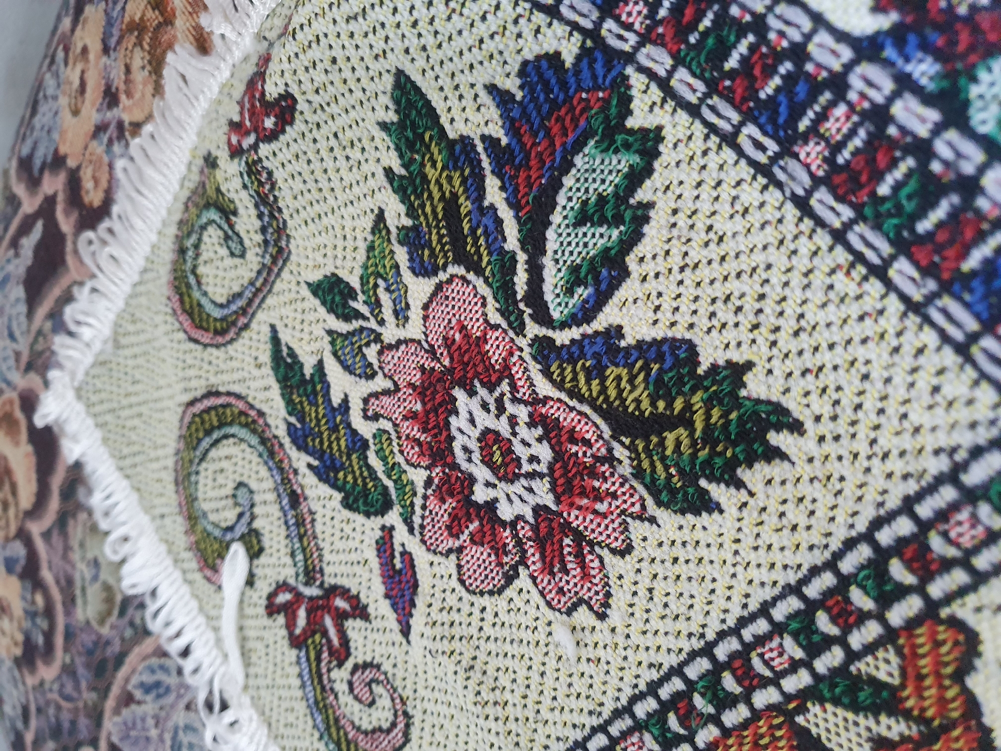 The image shows a close-up of a textured fabric with an embroidered floral design. The fabric is light beige with a woven pattern and features a central red and white flower surrounded by green leaves and blue accents. The design also includes intricate swirling patterns and a decorative border with geometric motifs in red, blue, green, and white. White fringe is visible along the top edge of the fabric. The background shows a blurred, darker fabric with a floral pattern. The image does not depict any specific location within Zubrești, Moldova, nor does it contain any people or actions.