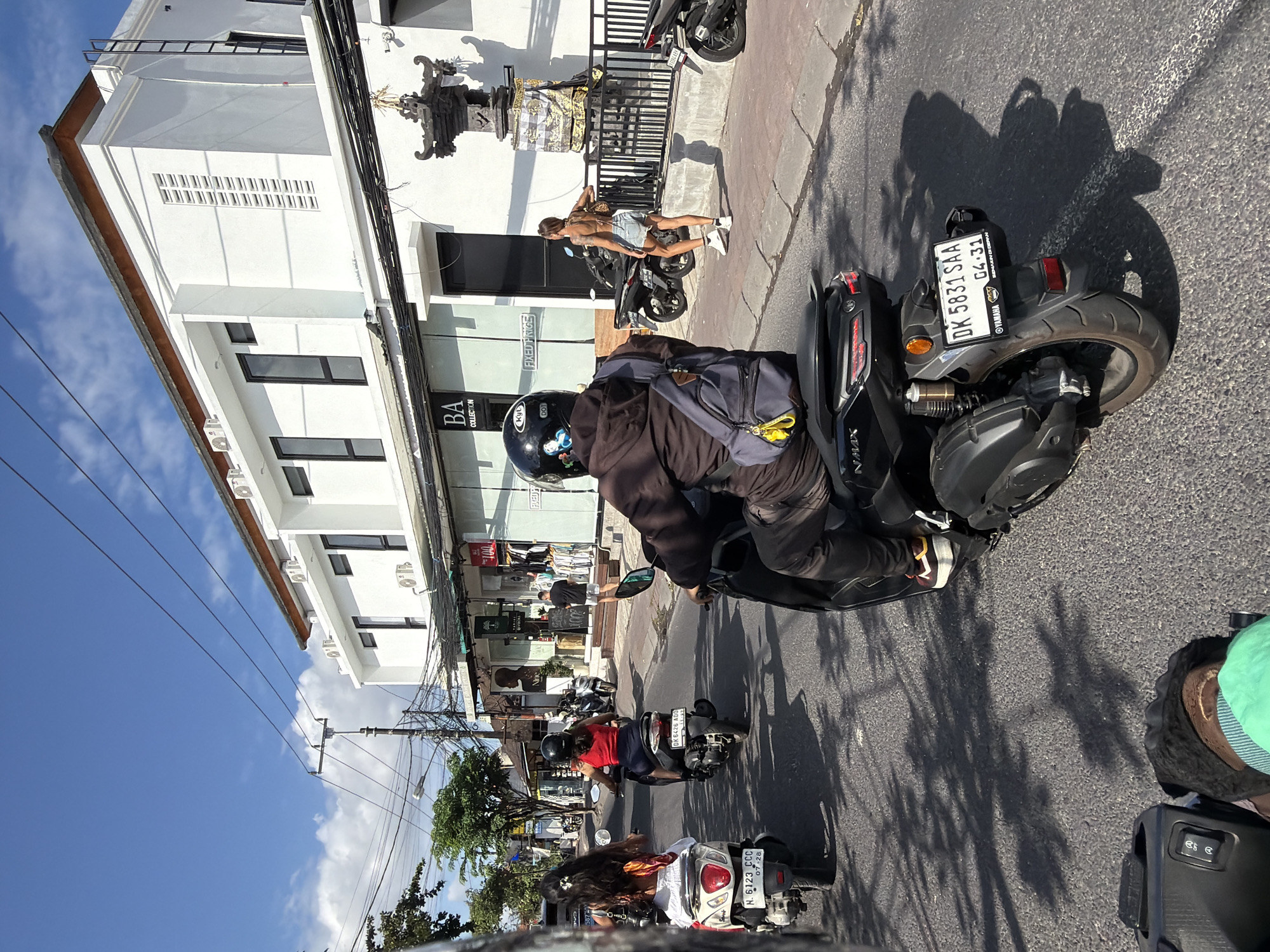 In North Kuta, Indonesia, a group of individuals rides motorcycles on a paved road under a clear blue sky with scattered clouds. A woman with long dark hair, wearing a white top and skirt with a colorful sarong, is on a white scooter. Next to her, a woman in a red tank top and dark pants rides a black scooter. Behind them, a person wearing a dark jacket, helmet, and backpack rides a black scooter. Further back, a person with a tattooed back, wearing a denim skirt and white sneakers, walks on the sidewalk. Buildings line the street, featuring white facades, dark windows, and awnings. Electrical wires crisscross overhead, supported by utility poles. Shadows of trees and objects are cast on the road. The scene depicts a typical day of motorcycle transportation in a commercial area.