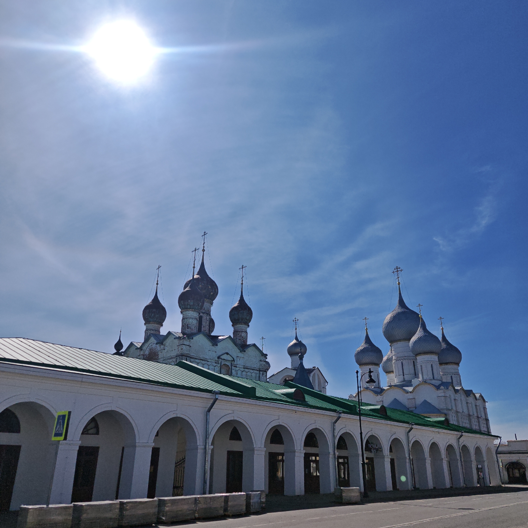 The image captures a low-angle view of a Russian Orthodox church complex and an adjacent arcade structure in Rostov, Russia. The sky is a clear blue with a bright sun at the top.
The church complex consists of multiple buildings with white walls and green roofs. The structures are topped with bulbous, metallic gray domes and darker, steeple-like domes, each adorned with crosses. The buildings appear to be old with some visible weathering.
A white arcade structure, featuring a series of arched openings, runs parallel to the street. A green roof covers the arcade, and the rectangular pillars supporting the arches cast shadows onto the walkway. A road runs along the side of the arcade. A street sign is present.