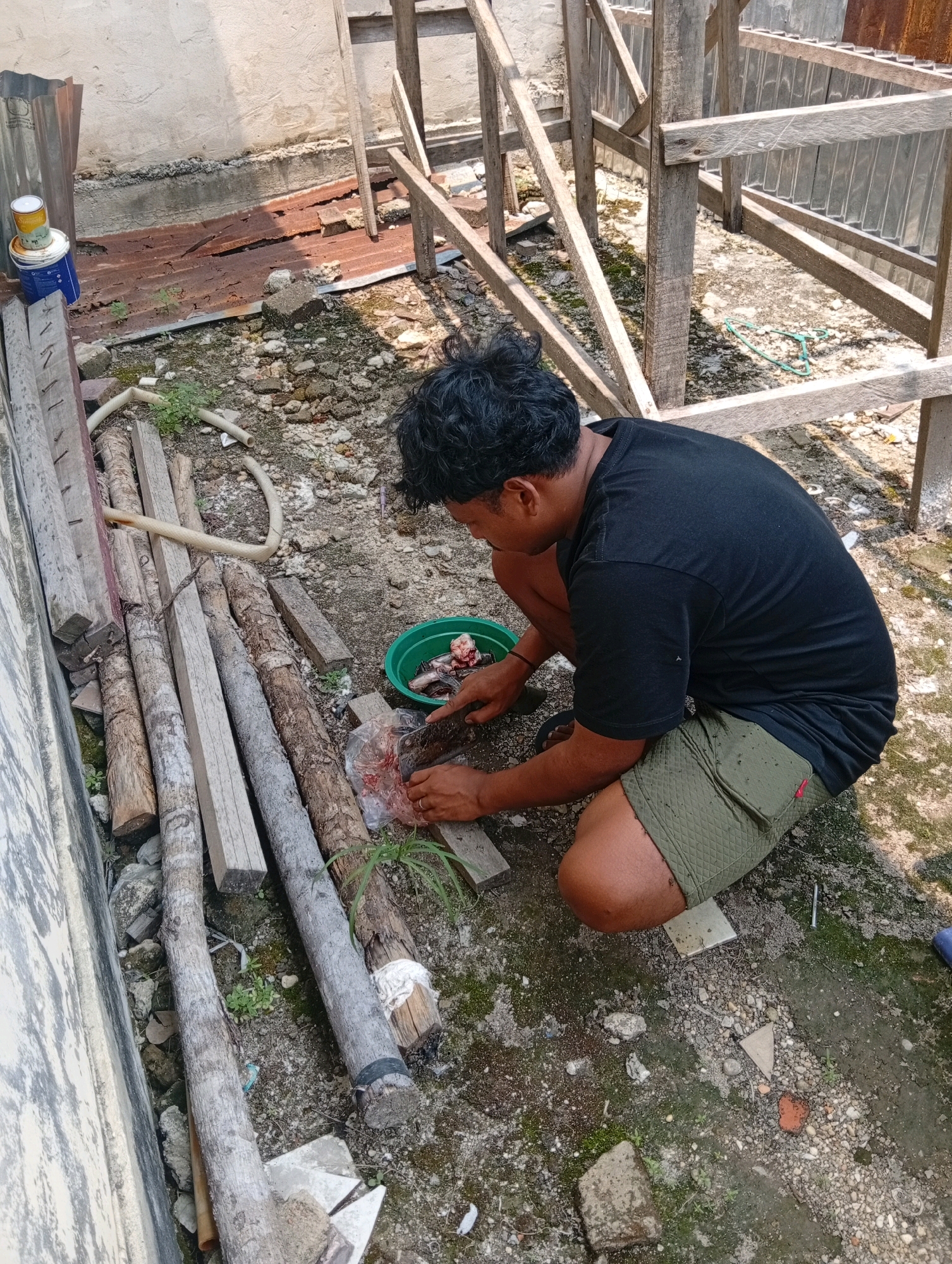 A medium shot captures a man in Kualu, Indonesia, crouched on the ground as he prepares fish outdoors. The man, wearing a black t-shirt and green shorts, is actively cleaning a fish with a knife. He is positioned to the right of a pile of logs and a green bowl containing more fish parts. The environment appears to be a construction or work area, with a partially constructed wooden structure visible in the background, along with scattered building materials like corrugated metal and concrete. The ground is covered with dirt, pebbles, and some sparse vegetation. The scene is illuminated by natural daylight, casting shadows on the ground.
