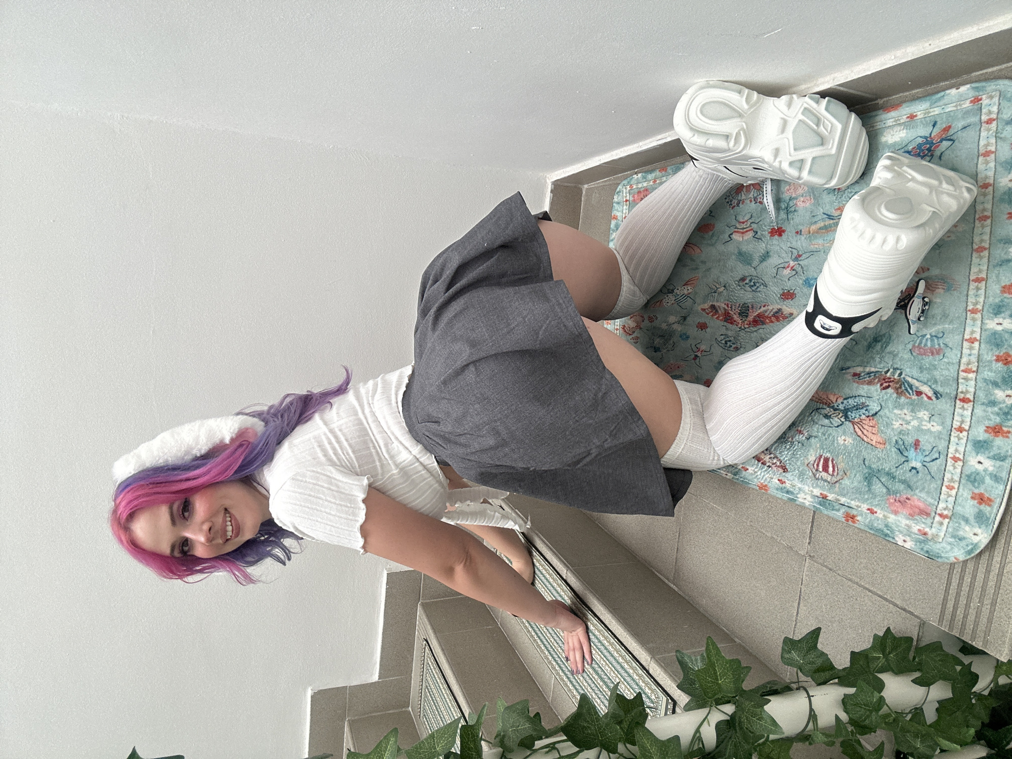 A young woman with pink and purple hair, wearing a white top, grey skirt, and white knee-high socks with chunky white sneakers, is lying on a staircase covered with a light blue patterned rug and faux ivy. She is smiling and looking directly at the camera. The location appears to be indoors, possibly a building in the City of Zagreb, Croatia, given the mention of the city. The walls are white, and the staircase is tiled.