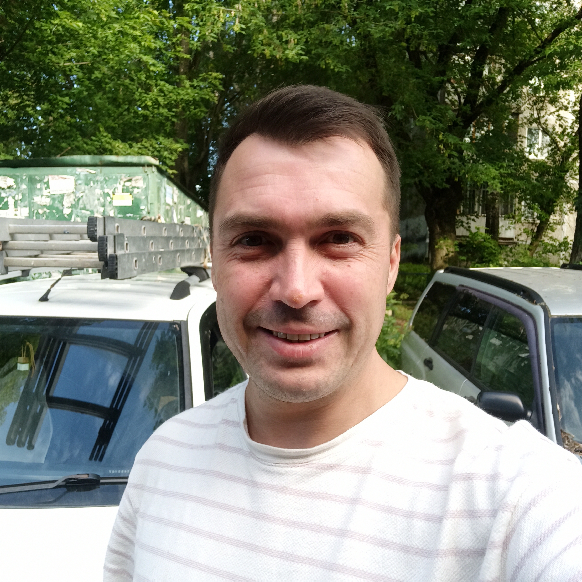 A man with short brown hair and stubble is pictured in a selfie. He is wearing a white t-shirt with thin, light pink stripes. He is smiling, showing his teeth. Behind him, a white car is visible, with a ladder secured to its roof. To the right of the white car is a silver car. In the background, trees with green foliage and a building with windows are present. The scene appears to be outdoors, possibly in a residential area or an industrial zone, given the presence of the cars and the dumpster. There are no visual cues to confirm the location as Nizhny Novgorod, Russia.