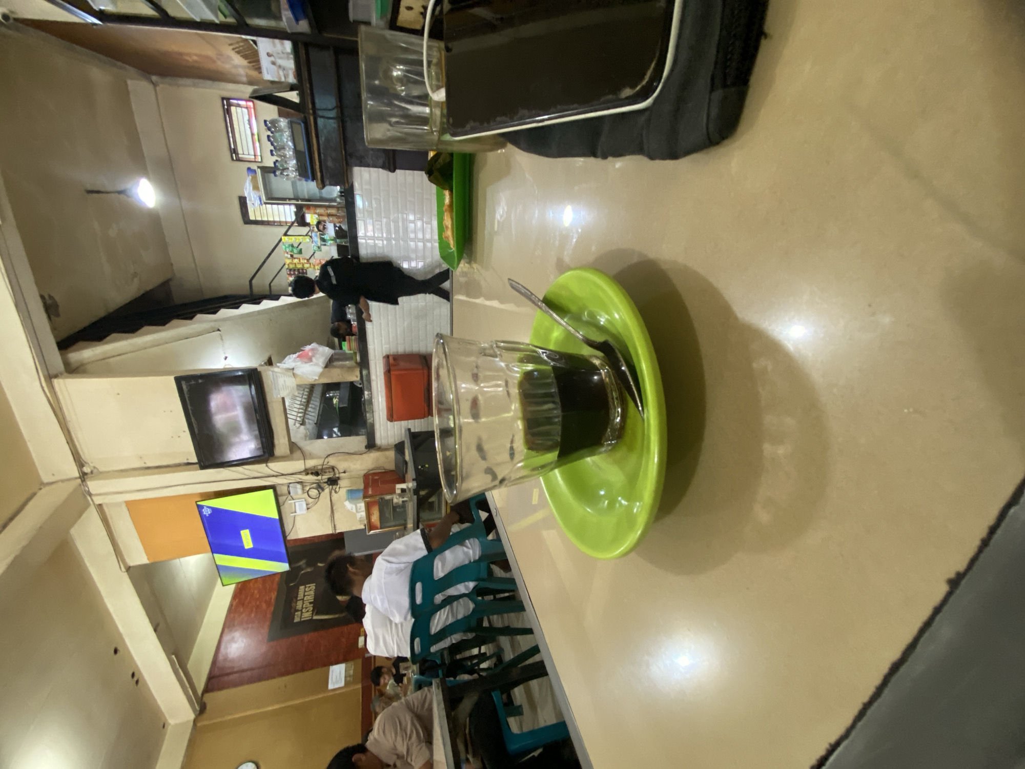 The image depicts the interior of a food establishment, likely a cafe or restaurant, in Lhokseumawe, Indonesia. In the foreground, a glass of dark liquid, presumed to be coffee, sits on a bright green saucer with a spoon. To the right, another glass and a black smartphone are visible on the table. The table itself is a light beige laminate surface.
In the middle ground, several people are seated at tables. Two individuals in white shirts are facing away from the camera, and another person in a light-colored shirt is partially visible on the left. Teal plastic chairs are arranged around the tables.
The background reveals a counter area where a person in a black uniform is standing. Behind the counter, shelves are stocked with bottles and other items. Two television screens are mounted on the wall; one displays a bright blue and yellow graphic, while the other shows a darker image. The overall environment suggests a casual dining setting with a functional, somewhat dated interior design, possibly illuminated by fluorescent ceiling lights. The context of Lhokseumawe, Indonesia, indicates a tropical urban setting.