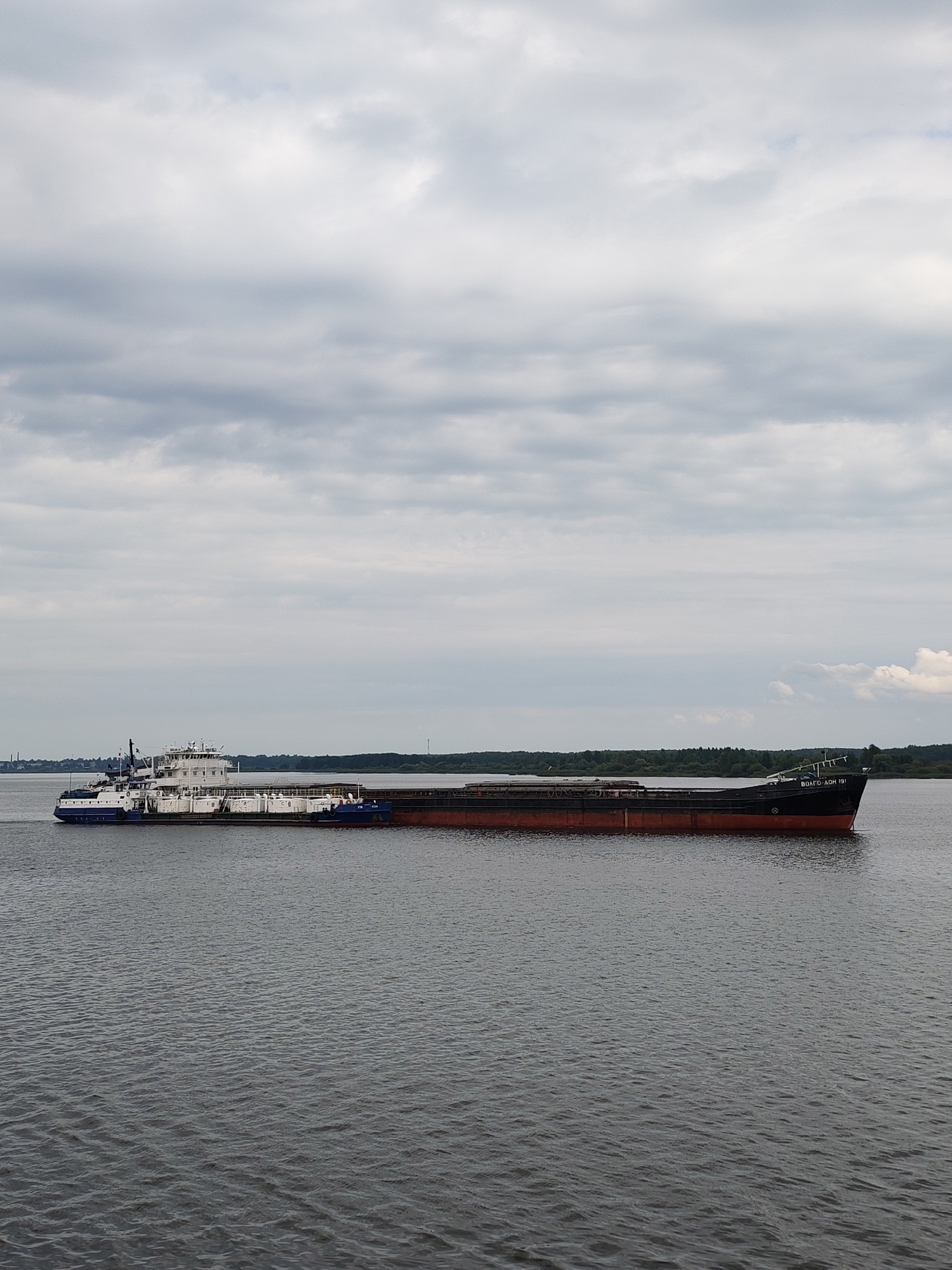 A towboat, the "VOLGO-DON 191", is pushing a barge on a wide, calm river under an overcast sky. The towboat is white and blue with a tall superstructure and appears to be heading towards the viewer's left. The barge is long and dark red with a black deck, and it is filled with cylindrical containers. The riverbanks in the distance are lined with a dense forest. The scene suggests a cargo transport operation on a waterway in Rybinsk, Russia.