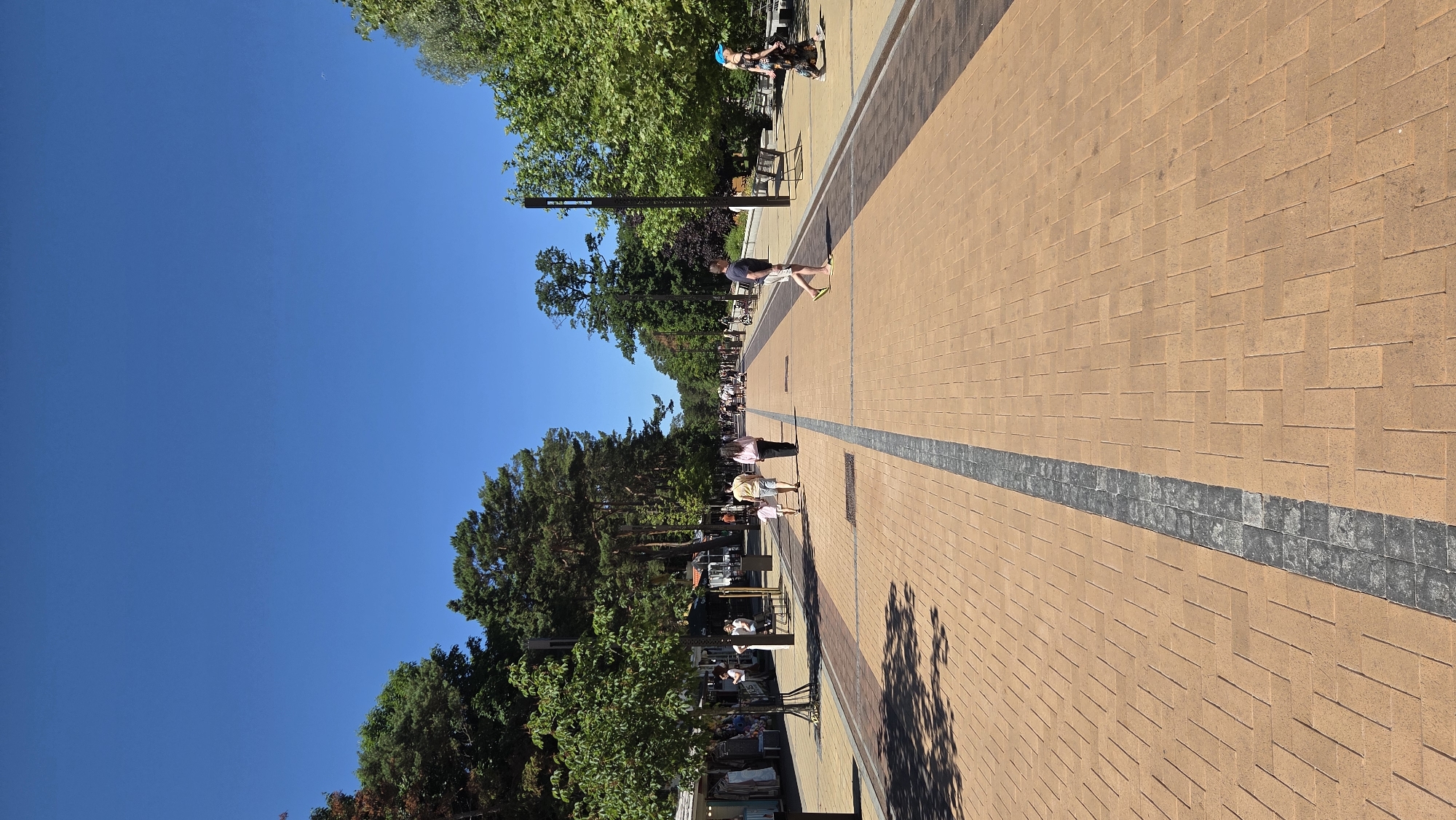 A street scene shows a wide pedestrian walkway paved with light brown bricks and a dark gray strip down the center. Trees line both sides of the walkway, with some buildings or stalls visible on the left side. Several people are walking in both directions, some with children. A man in a blue helmet is walking on the right side of the walkway. The sky is clear and blue.