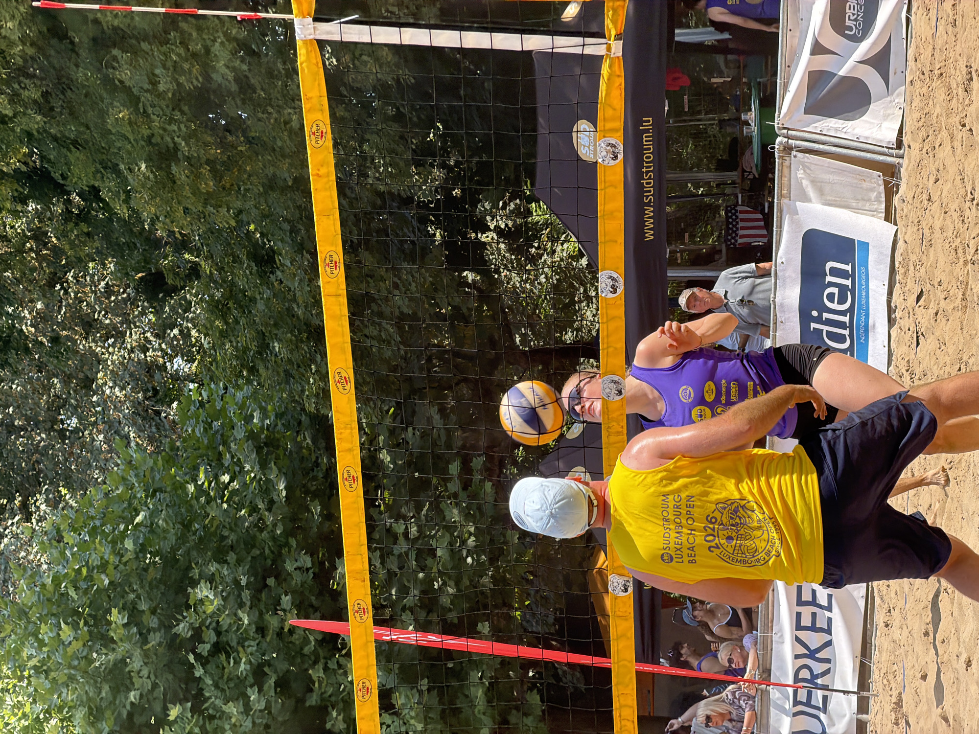 This image captures a beach volleyball match in Esch-sur-Alzette, Luxembourg. The foreground features two players, a man in a yellow tank top and a woman in a purple tank top, poised to play. The man, facing away from the camera, wears a light blue cap and dark shorts. The woman, looking up at the ball, wears black shorts. The volleyball is suspended mid-air above the net. The net itself is black with a yellow top band adorned with the "PITCHER" logo. Behind the players, a crowd is visible, some standing by a railing. Various banners are displayed, including one with "www.sudstroum.lu" and another with "dien INDÉPENDANT LUXEMBOURGEOIS." The environment is a sandy beach volleyball court, with lush green trees providing a backdrop. The lighting suggests a sunny day.
