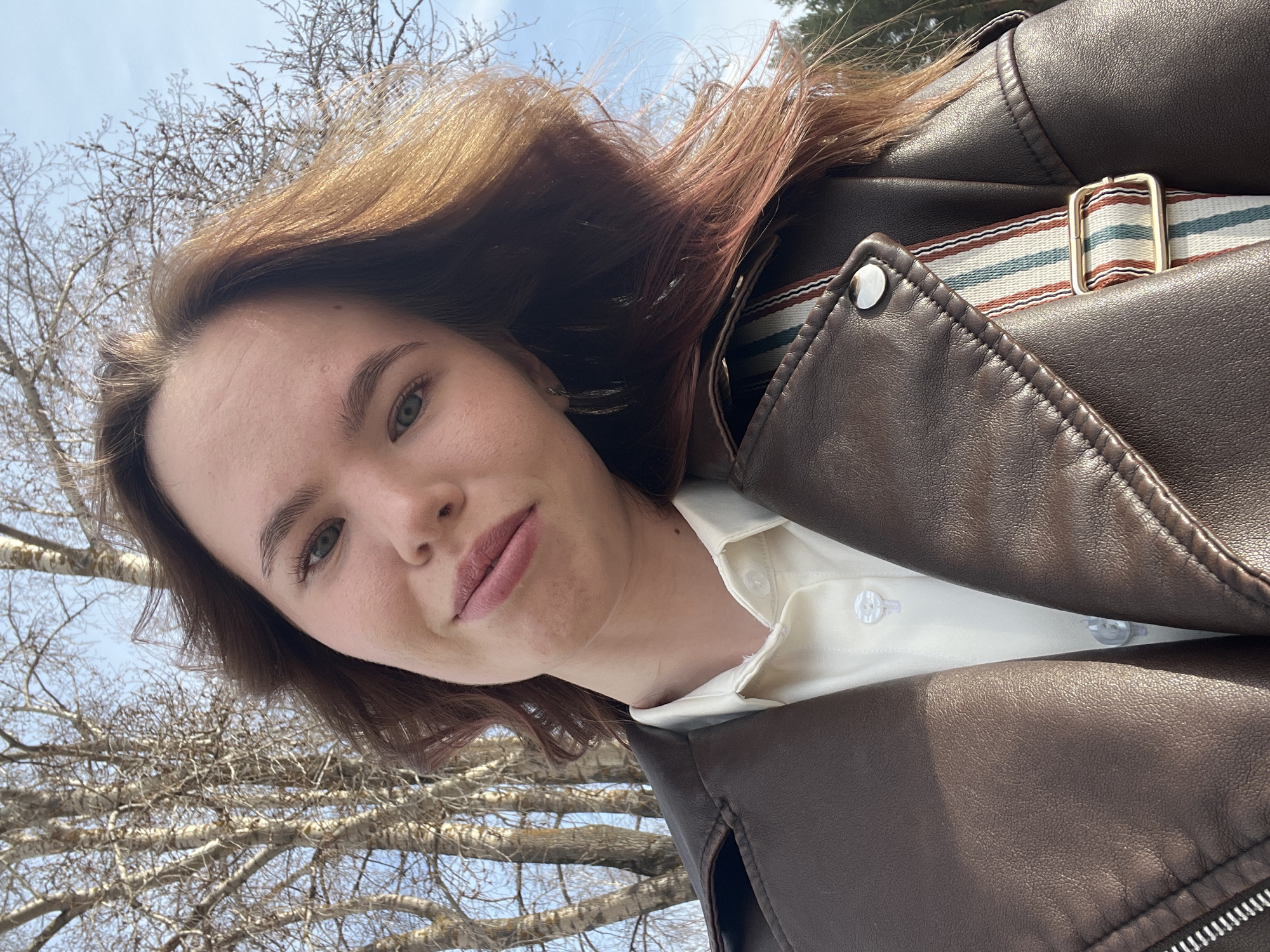 A photograph taken outdoors in daylight, presumably in Chaikovsky, Russia, depicts a woman wearing a brown leather jacket over a white collared shirt. The composition is taken from a low angle, with the woman's face in the upper portion of the frame, surrounded by bare tree branches and a patch of blue sky in the background. The texture of the leather jacket and the bark of the trees is visible, with a strap detail on the jacket. The woman has fair skin, light brown hair with hints of pink, and a neutral expression.
