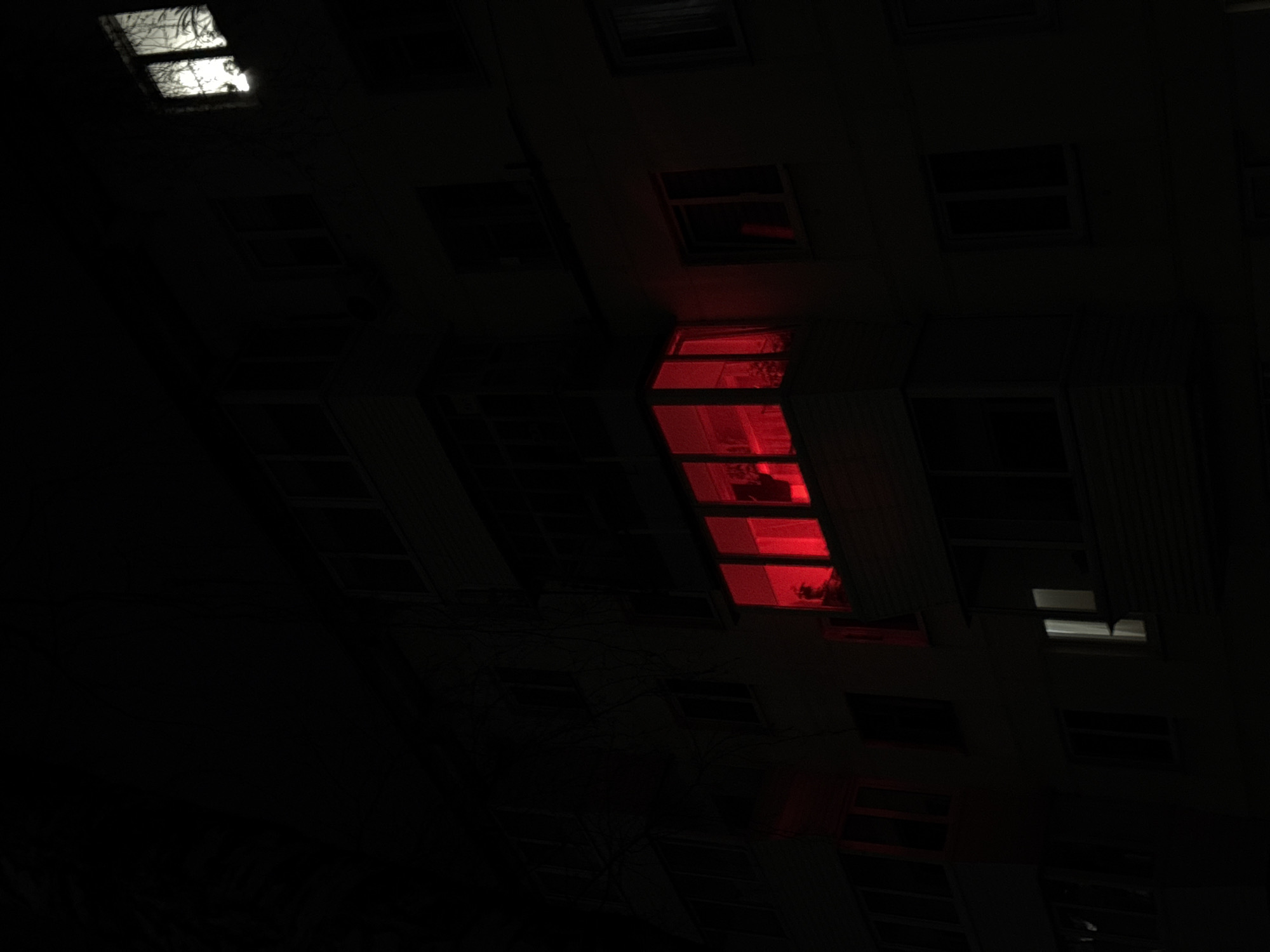 A night shot looking up at the corner of a building in Chaikovsky, Russia. Most windows are dark, but one vertical section of windows glows red. In the top left, one window emits a white light that is refracted by branches or debris. The scene is dark, with limited detail.