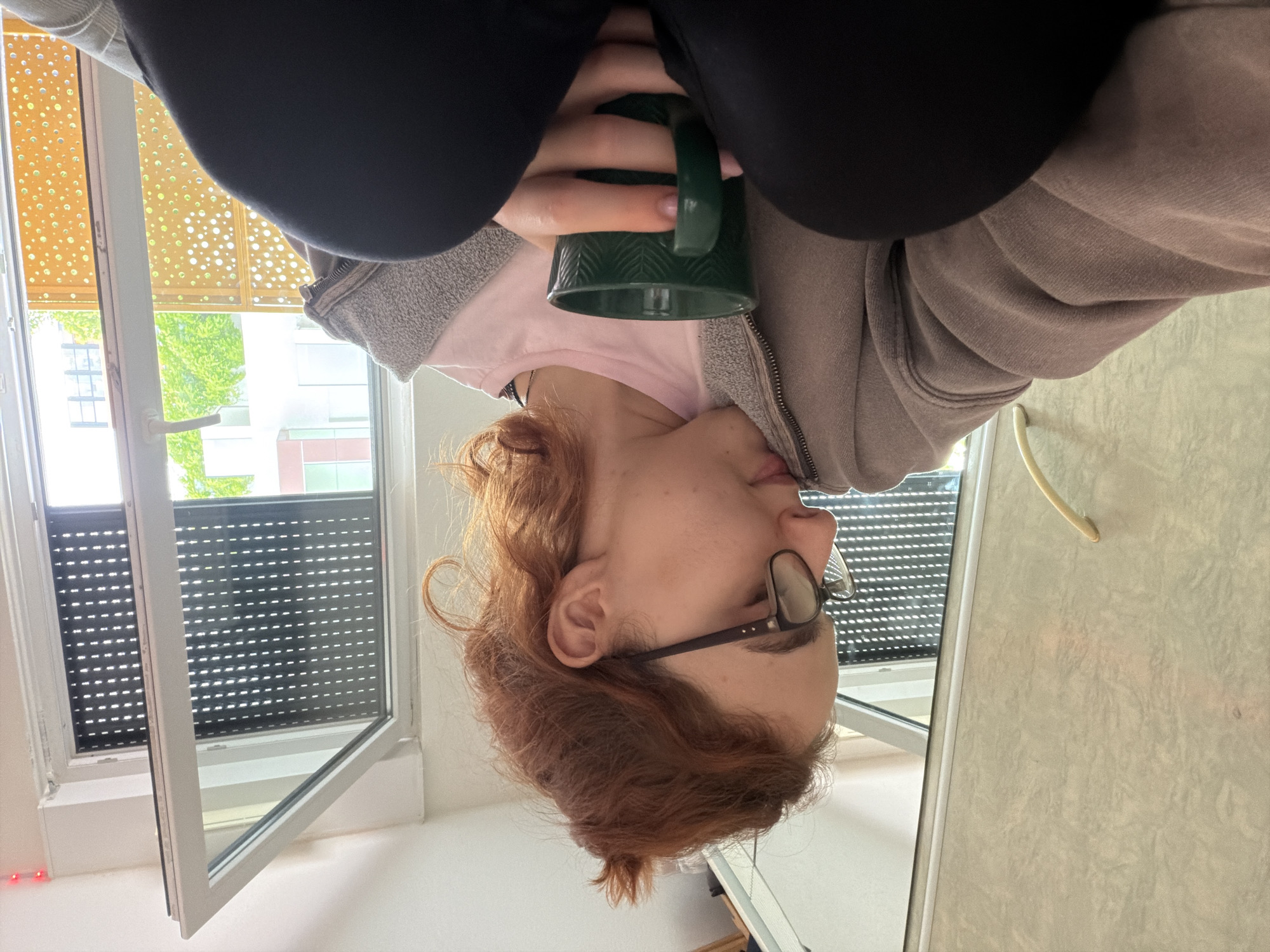 The image depicts a young woman with reddish-brown hair pulled up in a bun, wearing glasses and a light pink t-shirt layered under a gray zip-up hoodie. She is holding a dark green mug with a textured pattern and appears to be looking down, possibly in thought or while taking a drink. The scene is indoors, with a large window or glass door to her right, revealing a balcony with a patterned railing and some greenery outside. Reflections in the glass show parts of buildings. To the left of the woman is a portion of a closet or wardrobe with a light-colored, textured surface and a yellow handle. The overall setting suggests a casual, domestic environment, possibly an apartment in Strasbourg, France, given the architectural style visible through the window.