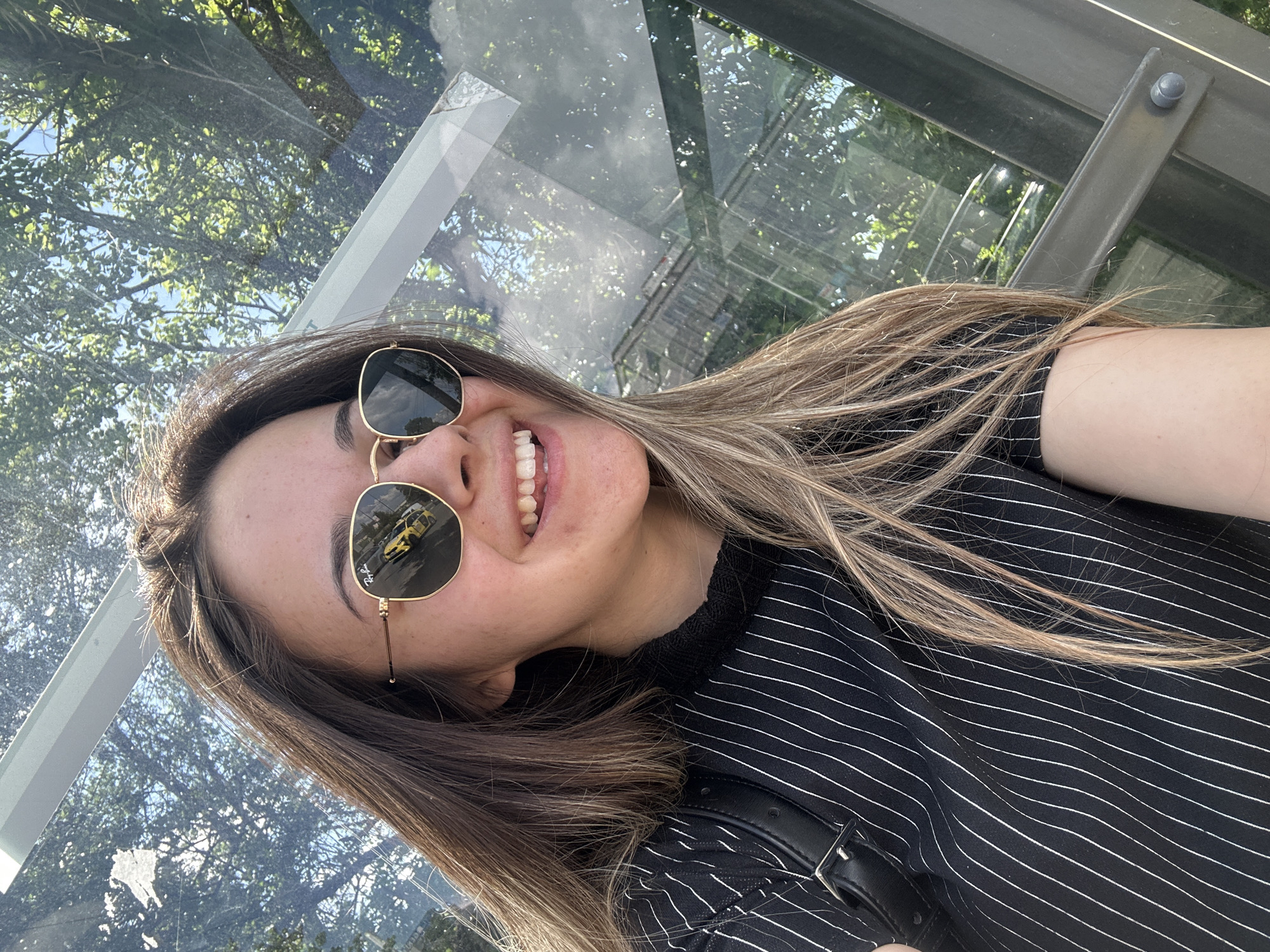 A young woman smiles while wearing sunglasses and a black and white striped shirt. The reflection in her sunglasses shows a yellow vehicle and what appears to be a building, likely in an urban setting. She seems to be standing at a bus or tram stop, based on the structure and glass panels behind her, which reflect trees. The image was likely taken during the day, under natural light. It is unknown if the location is in Sofia, Bulgaria as specified, because there are not enough details to confirm.