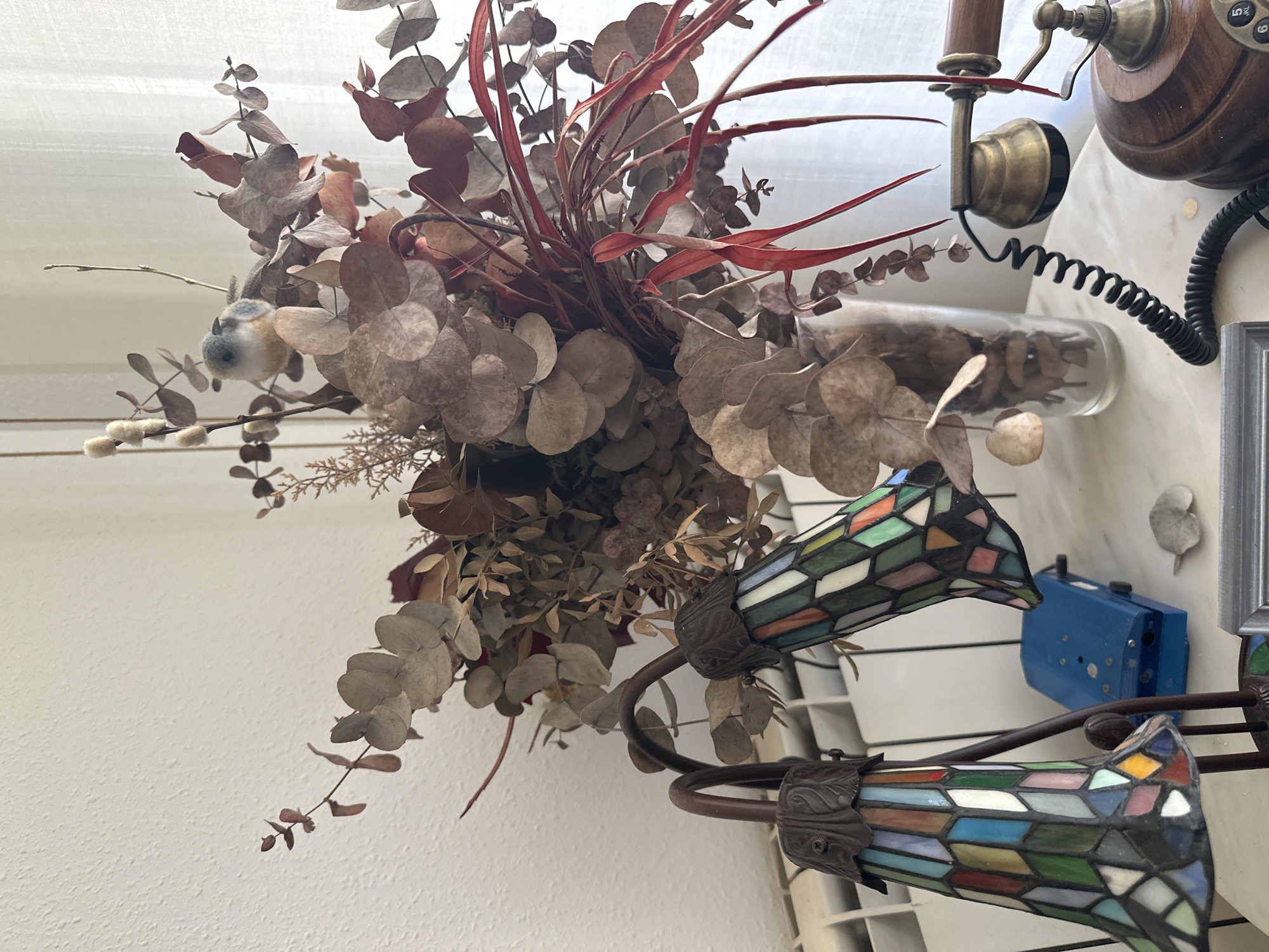The image depicts an indoor still life arrangement on a white surface, possibly a table or shelf, in a room located in Sant Pol de Mar, Spain. The scene features a decorative bouquet of dried eucalyptus and other foliage in a clear glass vase. Perched on the bouquet is a small, fuzzy bird figurine. To the left of the vase, a Tiffany-style lamp with a multi-colored stained-glass shade is visible. Behind the lamp, a white radiator runs horizontally. Adjacent to the lamp and vase, a vintage-style rotary telephone with a wooden base and handset rests on the surface. A coiled black cord connects the handset to the base. A small, bright blue object, possibly an electronic device, is situated between the lamp and the vase. The background consists of a textured white wall and a sheer white curtain. The lighting suggests it is daytime.