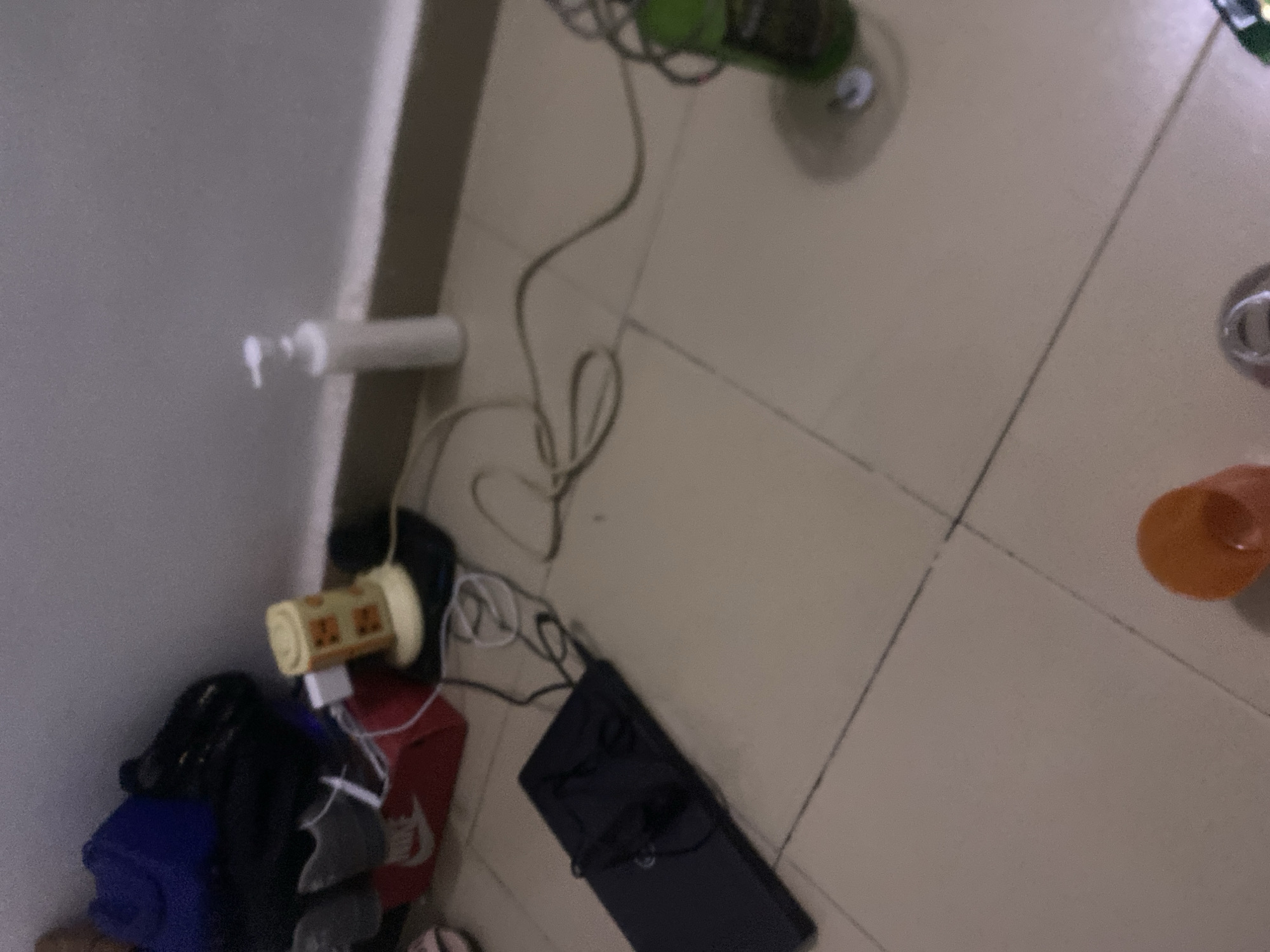 The image depicts a cluttered floor area in a room, possibly a bedroom or living space, located in Howayi, Nigeria. The floor is tiled in a neutral color. In the background, against a white wall, a dark blue bag and other dark items are piled up. A red shoebox with a Nike logo is visible, containing what appear to be shoes. Near the shoebox, a small, cylindrical object with a yellow and orange pattern sits on a black base. White charging cables are strewn across the floor, connected to various unseen devices. To the right, a white pump bottle of liquid is standing upright. Further to the right, a green bottle with a dark label and a white cap is placed on the floor, with a coiled cable nearby. In the foreground, a dark laptop is open and lying flat on the floor, with headphones resting on top of it. Another coiled cable is visible next to the laptop. In the bottom portion of the image, an orange cup or container is partially visible, along with what appears to be a metal ring or lid. The overall scene suggests a lived-in space with items scattered around, possibly indicating recent use or disorganization.