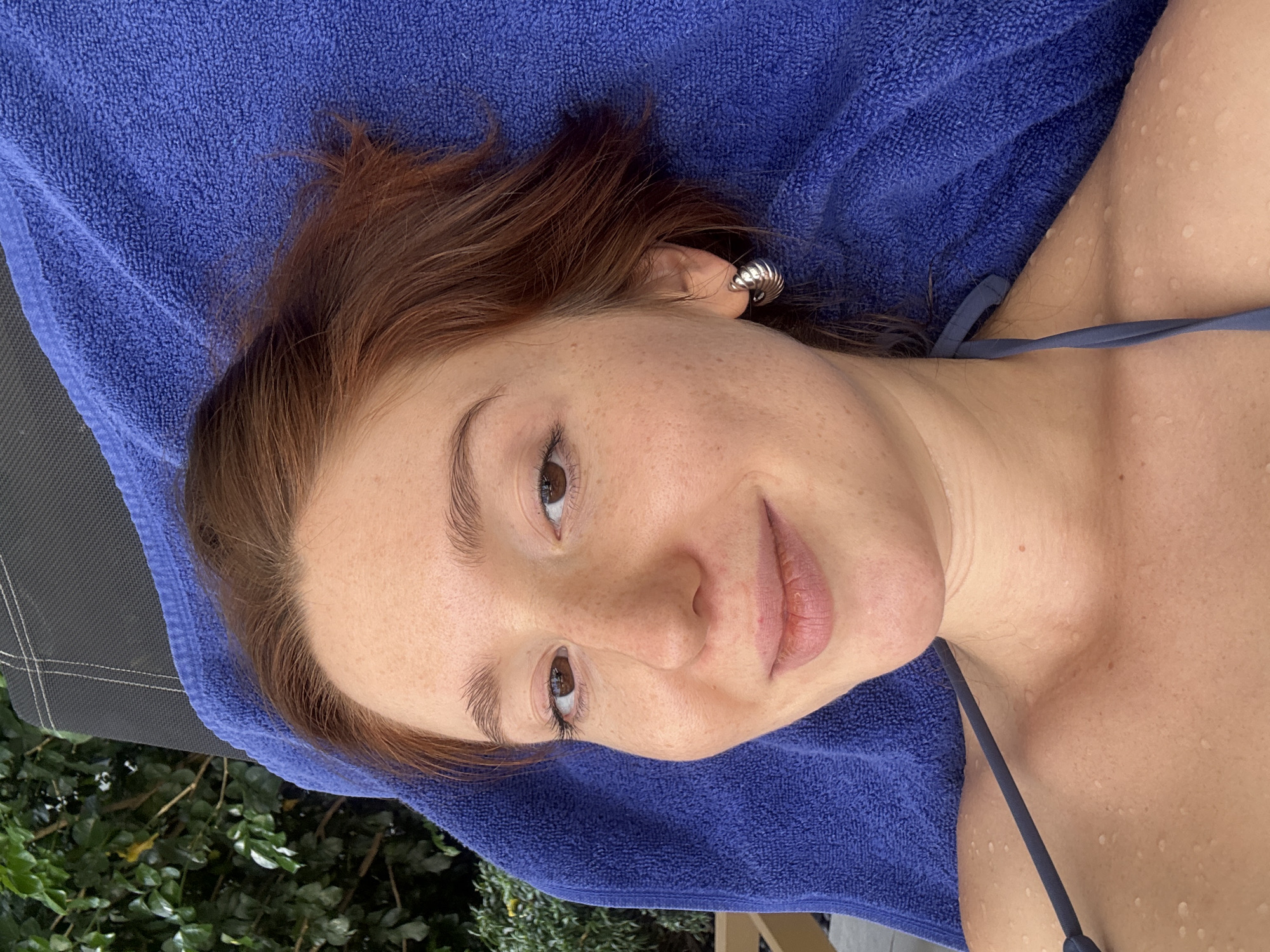 A young, fair-skinned woman with freckles, light brown eyes, and reddish-brown hair lies on a blue towel. She is wearing a dark blue swimsuit. Her skin appears slightly wet with water droplets. An earring is visible in her left ear. The edge of what seems to be a dark-colored lounge chair is visible at the bottom left. In the lower-left corner, green foliage suggests an outdoor setting. The location is indicated as Choeng Thale, Thailand, although this is not evident from the image itself.