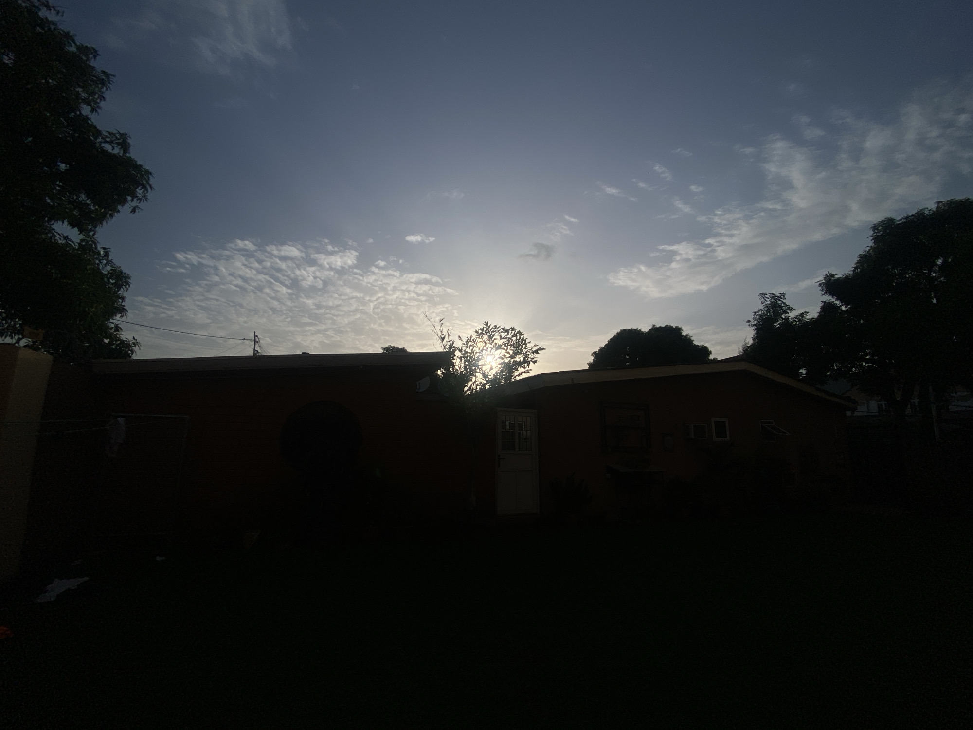 In Barden Yana, Nigeria, the image depicts a residential building silhouetted against a dusky sky. The sun, partially obscured by trees, casts a soft glow through the scattered clouds, creating a serene atmosphere. The building, painted in warm earth tones, features a white door and several windows, one of which is slightly ajar. Lush trees frame the scene, their dark foliage contrasting with the lighter sky. The foreground is a dark expanse of lawn, hinting at evening or early morning light.
