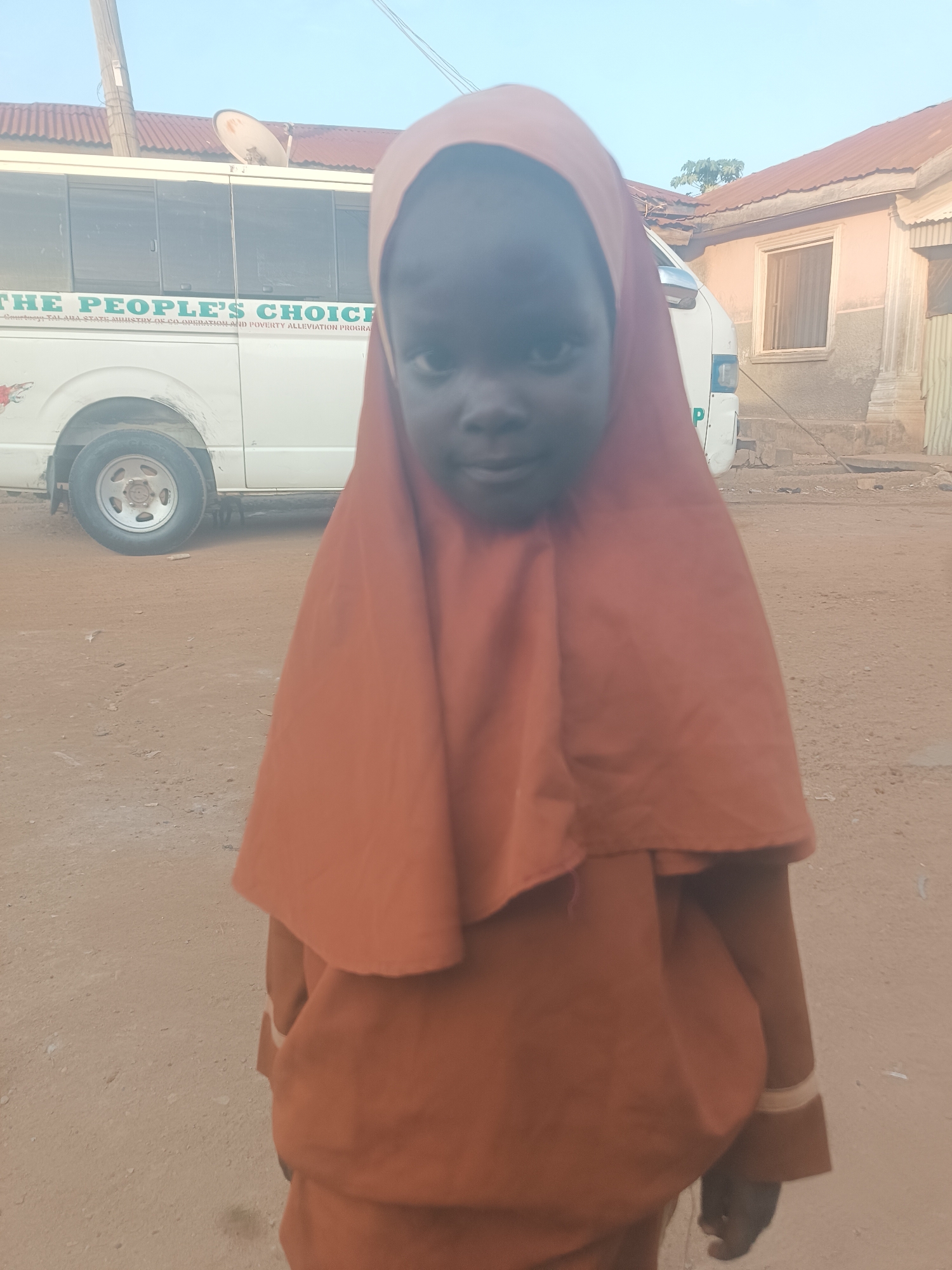 A young girl wearing an orange hijab and matching outfit stands in front of a white bus with "THE PEOPLE'S CHOICE" written on its side. The bus also has text indicating it is part of a "TARIA STATE MINISTRY OF CO OPERATION AND POVERTY ALLEVIATION PROGRAM." The girl's face is visible, looking towards the camera. The ground is dusty and sandy, consistent with an outdoor environment in Wukari, Nigeria. Buildings with corrugated iron roofs are visible in the background. The scene appears to be an everyday street view.